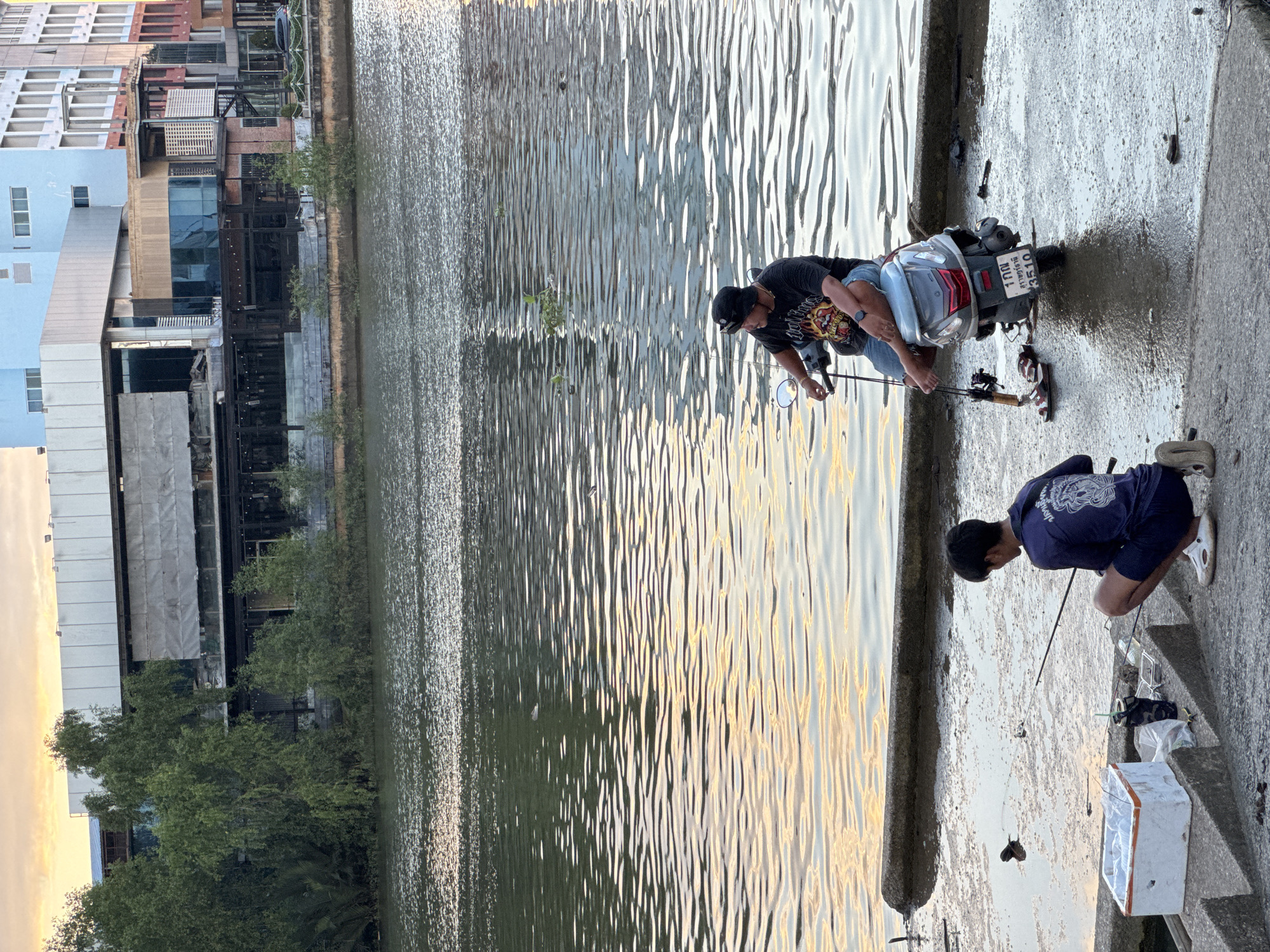 In Surat Thani City Municipality, Thailand, two individuals are engaged in fishing by the waterfront. A man, seated on a scooter parked on the wet concrete embankment, is holding a fishing rod. Beside him, a younger person kneels on the embankment, also with a fishing rod, facing the water. In the foreground, a white styrofoam cooler box and other fishing accessories are placed on concrete steps leading down to the water's edge. The water reflects the soft colors of the sunset sky and is disturbed by ripples. Behind the embankment, a multi-story building with a modern architectural design is visible, surrounded by trees and other structures, suggesting an urban setting.