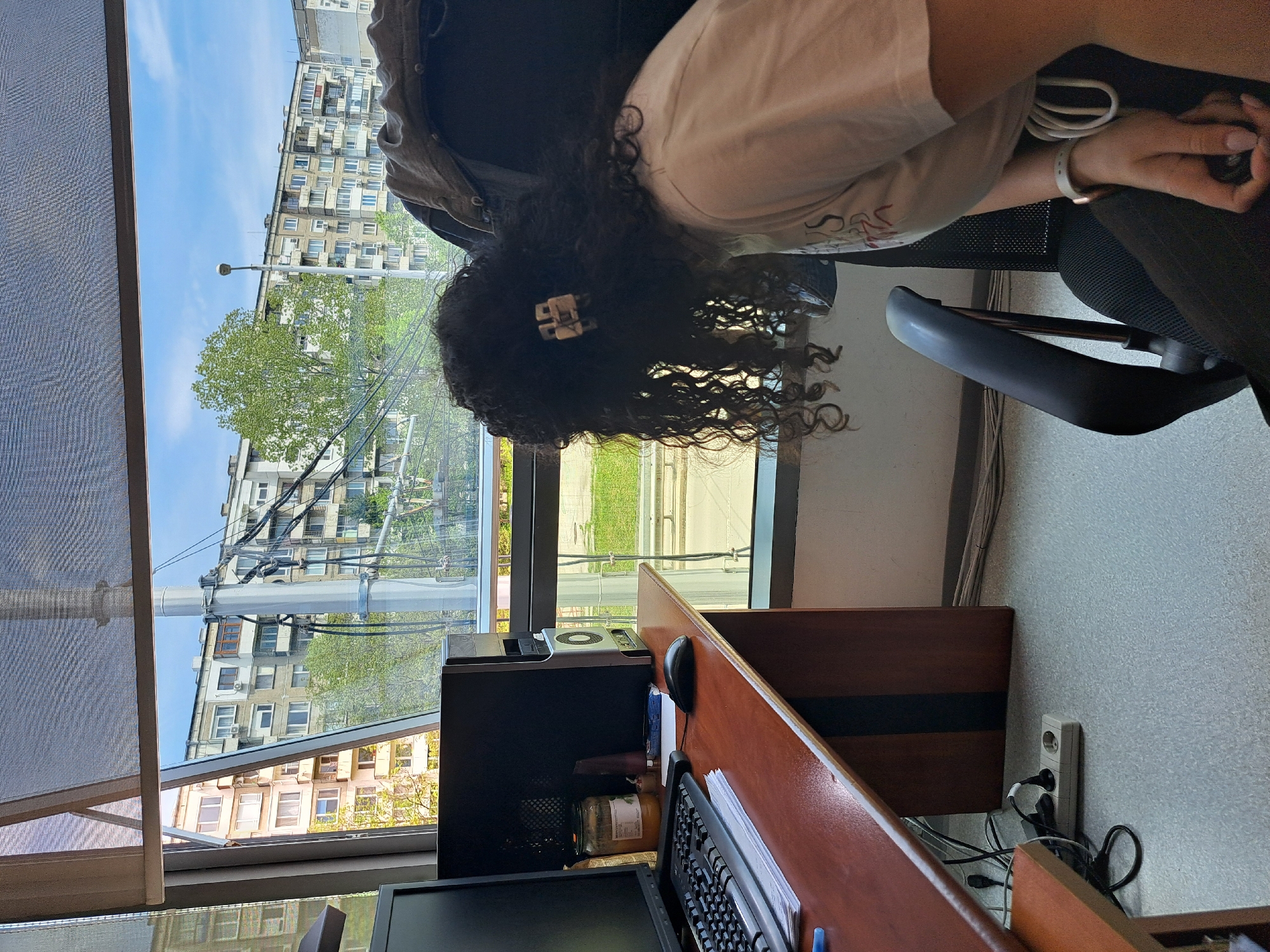 The image captures a vertical view of a person leaning over an office desk, with a window revealing an urban landscape.
**Subjects:**
The primary subject is a person, seen from the back and side, with long, dark, curly hair held back by a light-colored, rectangular hair clip. They are wearing a light-colored t-shirt with some indistinct red/orange stylized writing and dark pants. The person is seated at a wooden desk or cubicle, leaning forward with hands clasped together, wearing a white wristband on their left wrist.
On the desk are a black computer monitor, a black desktop computer tower, a keyboard, and a mouse. A glass jar with a label is placed next to the computer tower. A grey office chair is partially visible.
Through a large window, a bright blue sky with scattered clouds is visible. Outside, a tall, multi-story residential building with numerous windows and balconies stands prominently. Green trees, utility poles, and wires are also part of the outdoor scene. Part of the window on the left is covered by a grey, textured blind.
**Activity or event taking place:**
The person appears to be focused on something on the desk or computer, or perhaps resting their head in their hands while seated at the workstation.
**Notable details:**
The time of day is daytime, indicated by the bright natural light and clear blue sky. The exterior architecture suggests a dense urban environment. An electrical outlet strip with several plugs is visible on the wall next to the desk. The prominent curly hair and the hair clip are distinctive features of the person.
**Visible text:**
The word "NATURAL" is partially legible on the label of the jar next to the computer tower. Text on the person's shirt is too blurry to be read.