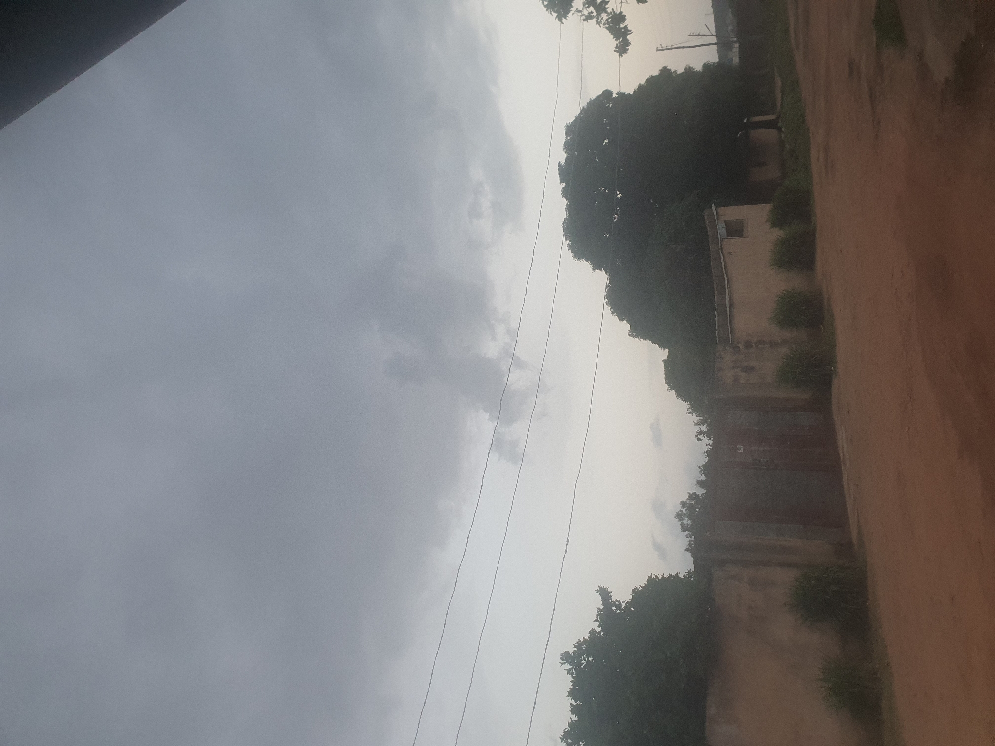 This image depicts a scene in Jos, Nigeria, likely during the late afternoon or early evening given the soft, diffused light. The sky is overcast, suggesting cloudy or potentially rainy weather. The composition is oriented vertically, with the right side of the image showing a dirt road or path and a wall with a large, weathered metal gate. Trees and lush green vegetation flank the wall and extend into the background.
Further back, a low-rise building with a simple design is visible, partially obscured by foliage. A single, small window is discernible on its facade. Another, larger structure with a tin roof is also present. Power lines crisscross the sky, a common sight in urban and semi-urban areas. The overall impression is one of a somewhat rustic or older neighborhood. There is no visible text in the image. The scene appears calm and quiet, with no people or animals actively present.