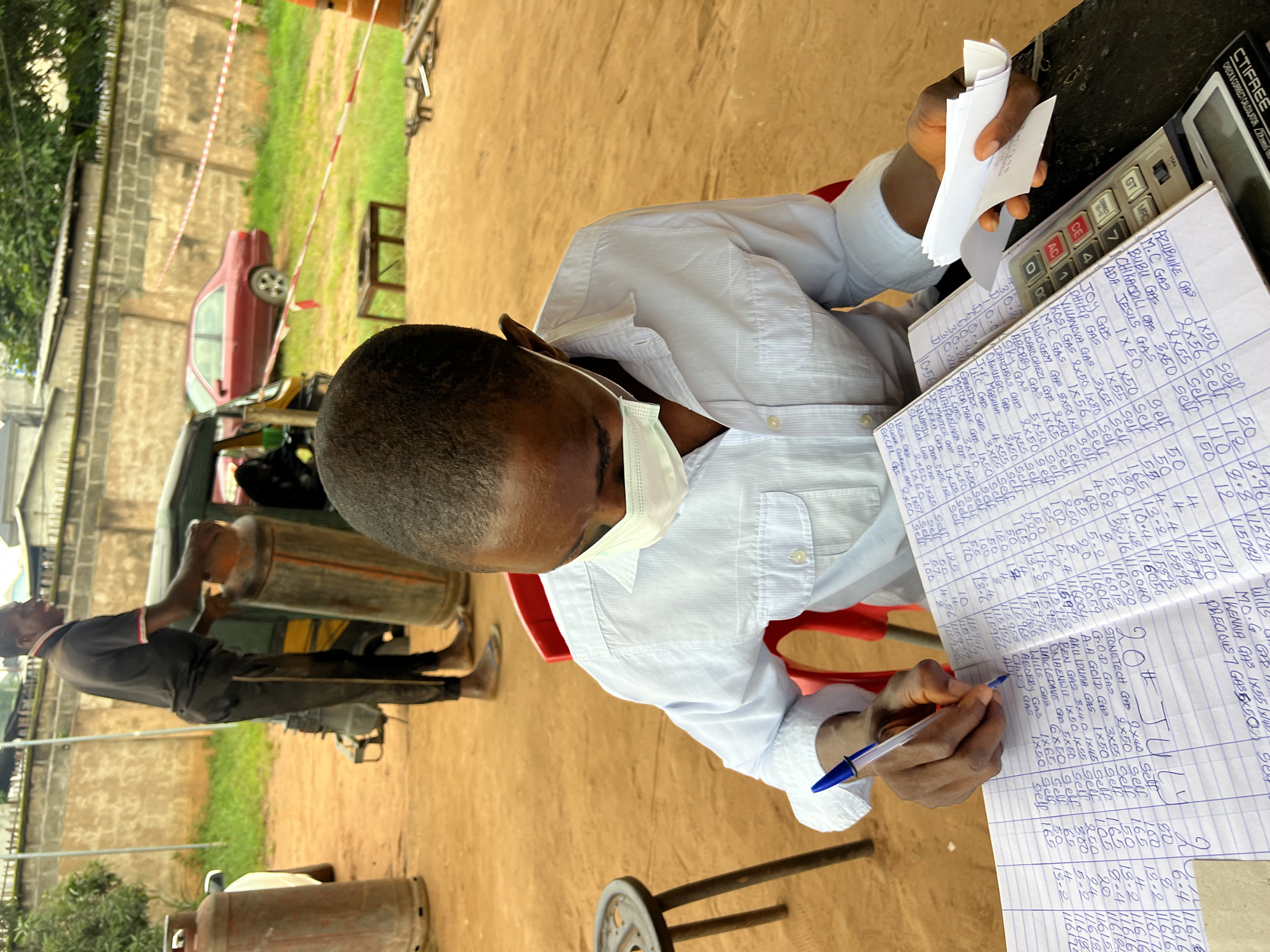 In Nkpor, Nigeria, a young man wearing a light blue striped dress shirt and a white face mask is seated at a table, diligently writing in a ledger. His right hand holds a blue and silver pen, poised over the open notebook. To his right, a calculator rests on the table, and he holds a small stack of white receipts in his left hand. The ledger contains numerous entries with columns for dates, names, quantities, and amounts. Behind him, another man, wearing a black t-shirt and black pants, is visible in the mid-ground, standing next to a large gas cylinder and appearing to be engaged in some activity related to it, possibly loading it onto a three-wheeled vehicle that is partially obscured. Gas cylinders and other objects are scattered around the outdoor area. The ground is covered in reddish-brown earth, and in the background, a red car, a wall, and some vegetation are visible, suggesting a semi-urban or commercial environment.