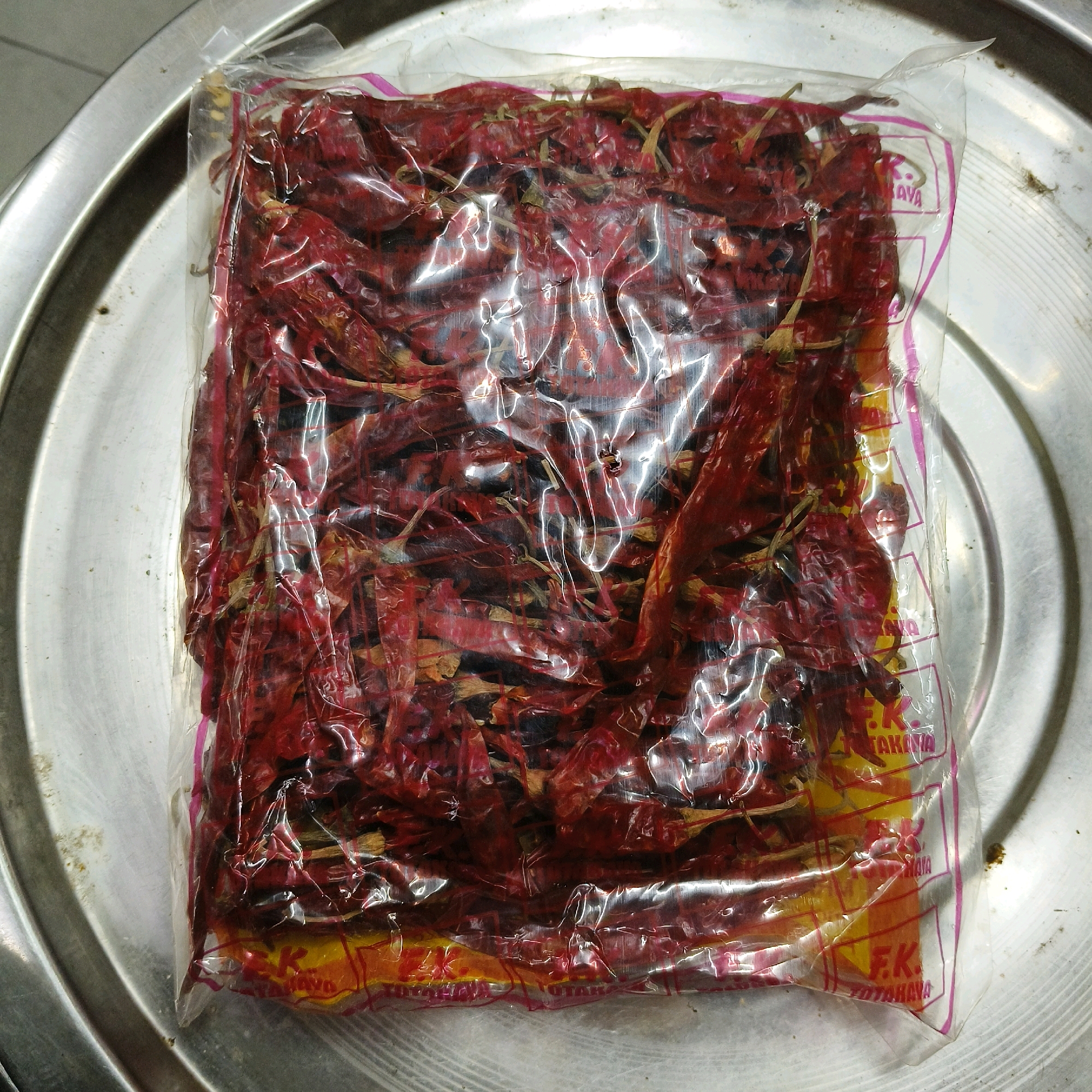 A plastic bag filled with dried red chilies is placed on a metallic circular tray. The plastic bag is partially transparent, revealing the contents inside. The chilies are a deep red color and appear to be whole, with some stems still attached. The bag has some text and graphics printed on it, including what appears to be "F.K." and "TOTAKAYA" in red and yellow. The metallic tray has a textured surface and a rim. The background is out of focus but appears to be a flat surface. There are no people or specific actions depicted in the image. The setting is indoors, likely a kitchen or storage area, given the presence of dried chilies and a tray. It is not possible to determine the specific location in Karimnagar, India from the image alone.