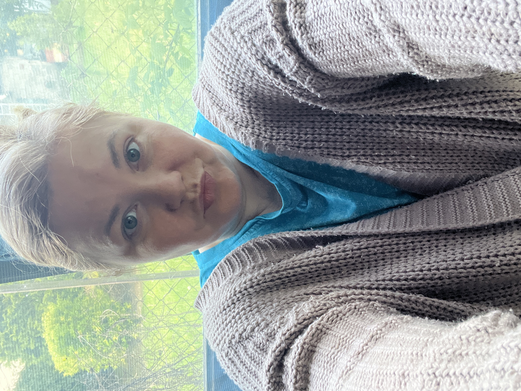 A woman with fair skin, blond hair, and blue eyes is centered in the frame. She wears a blue shirt under a large, knitted, brown cardigan. She looks at the camera with a slight smirk. Behind her is a window with a view of greenery outside, possibly a garden or park. The location is not identifiable as Vesyoloye, Russia based on the image alone.
