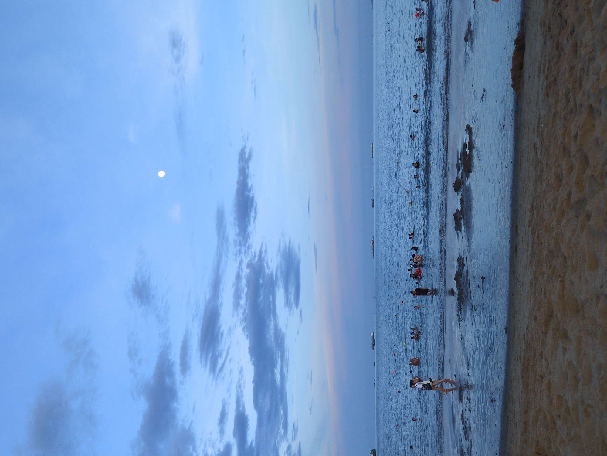This is a vertical, full-shot photograph of a beach scene at dusk in An Nhơn, Vietnam. The sky is a gradient of pale blue to a soft pink, with scattered clouds. A small, bright full moon is visible in the upper right quadrant of the sky. The ocean is calm and filled with numerous people swimming and wading. In the distance, several cargo ships are visible on the horizon. The foreground of the image is a sandy beach with some wet sand and small pools of water reflecting the sky. There is a solitary woman walking on the wet sand near the water's edge, carrying a bag. The overall mood is peaceful and serene, capturing a moment of leisure at the end of the day.