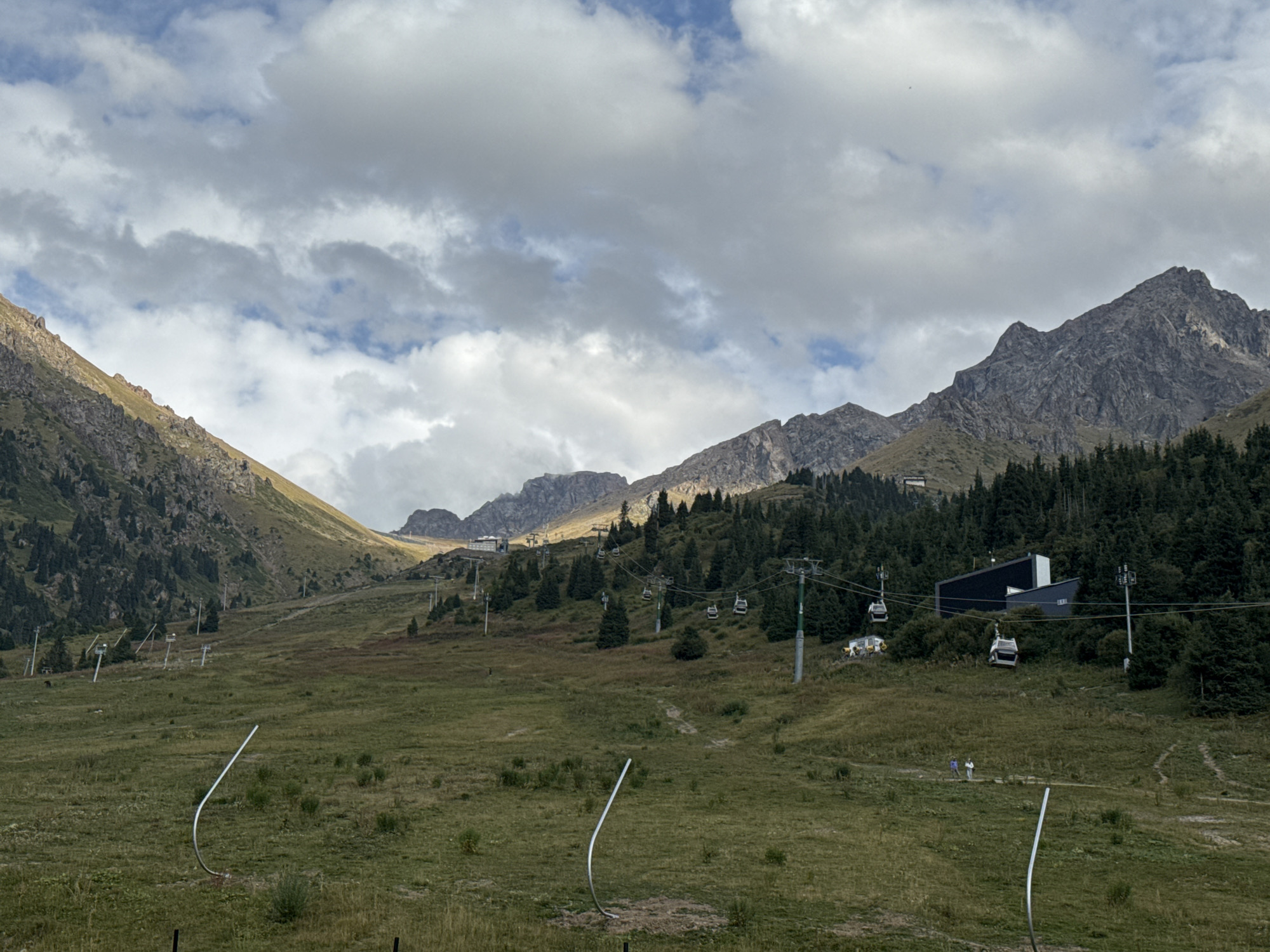 The image shows a view of the Medeu high-mountain ice skating rink and the Shymbulak ski resort area near Almaty, Kazakhstan. In the foreground, a grassy meadow is visible with several bent metal poles. In the middle ground, a ski lift operates, with several cabins suspended from cables that traverse the mountainous terrain. The slopes are covered in green grass and interspersed with evergreen trees. In the background, the rugged, rocky peaks of the Tian Shan mountains rise majestically under a cloudy sky. A modern building, likely part of the resort facilities, is situated on the slope. Two small figures, appearing to be people, are walking on a trail in the middle ground.