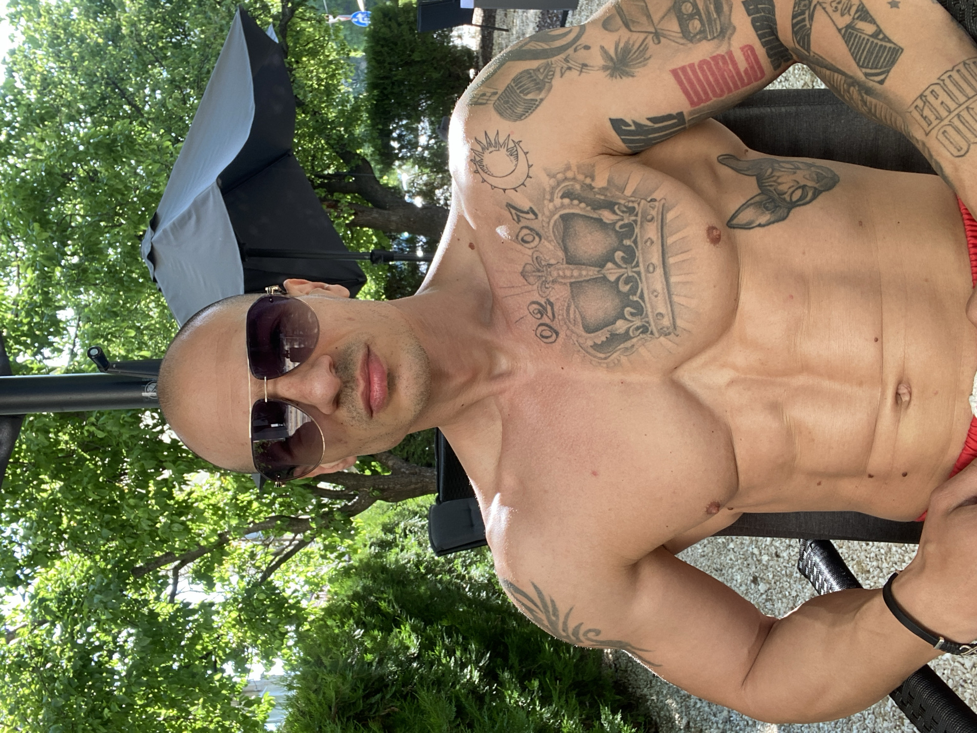 A shirtless man is lying back on a chair, with a black wristband on his left wrist. He is wearing sunglasses and has multiple tattoos covering his upper body, including tattoos of a crown and the word "WORLD". He is in an outdoor setting with green trees and foliage visible in the background. There is no information to determine if the photo was taken in Pleven, Bulgaria.