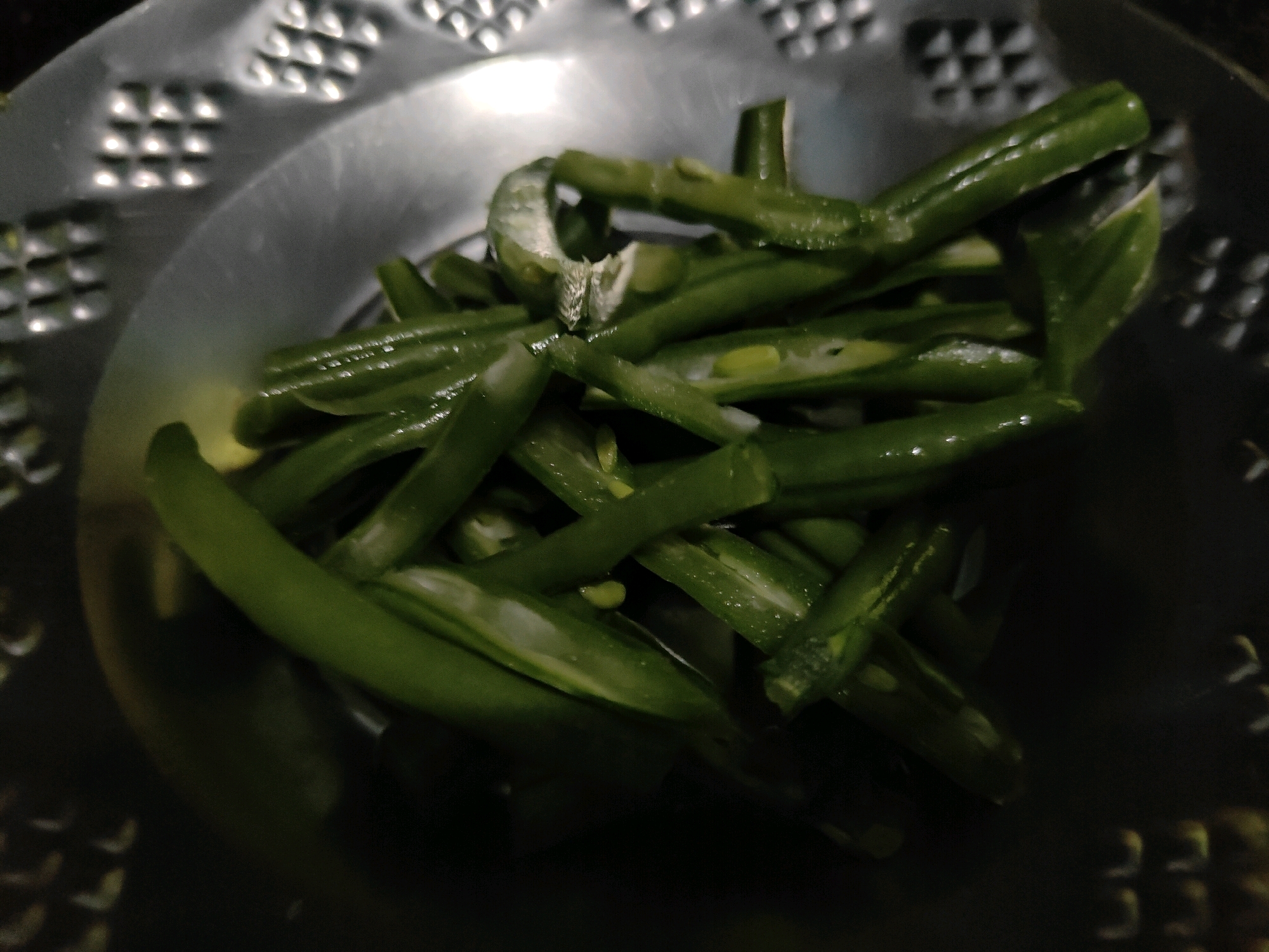 A close-up, overhead shot shows a pile of chopped green beans in a metallic colander. The beans are a vibrant green, some appearing shiny and wet. The colander has a patterned interior with raised dots arranged in rows. The background is dark and out of focus. There are no people or animals present. The image is of food preparation, likely in a kitchen.