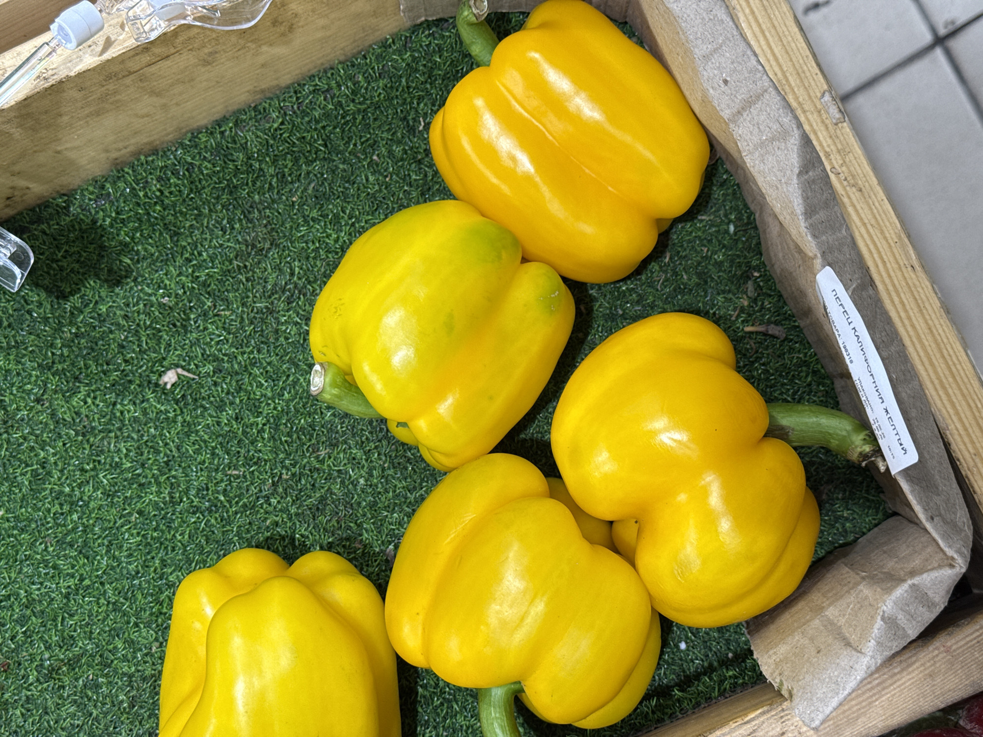 A top-down, close-up shot shows a wooden crate filled with bright yellow bell peppers resting on a green artificial turf surface. Six bell peppers are visible, some clustered together, with their green stems protruding. A white paper label with Russian text is partially visible at the bottom of the crate. The crate is positioned on a grey tiled floor.
