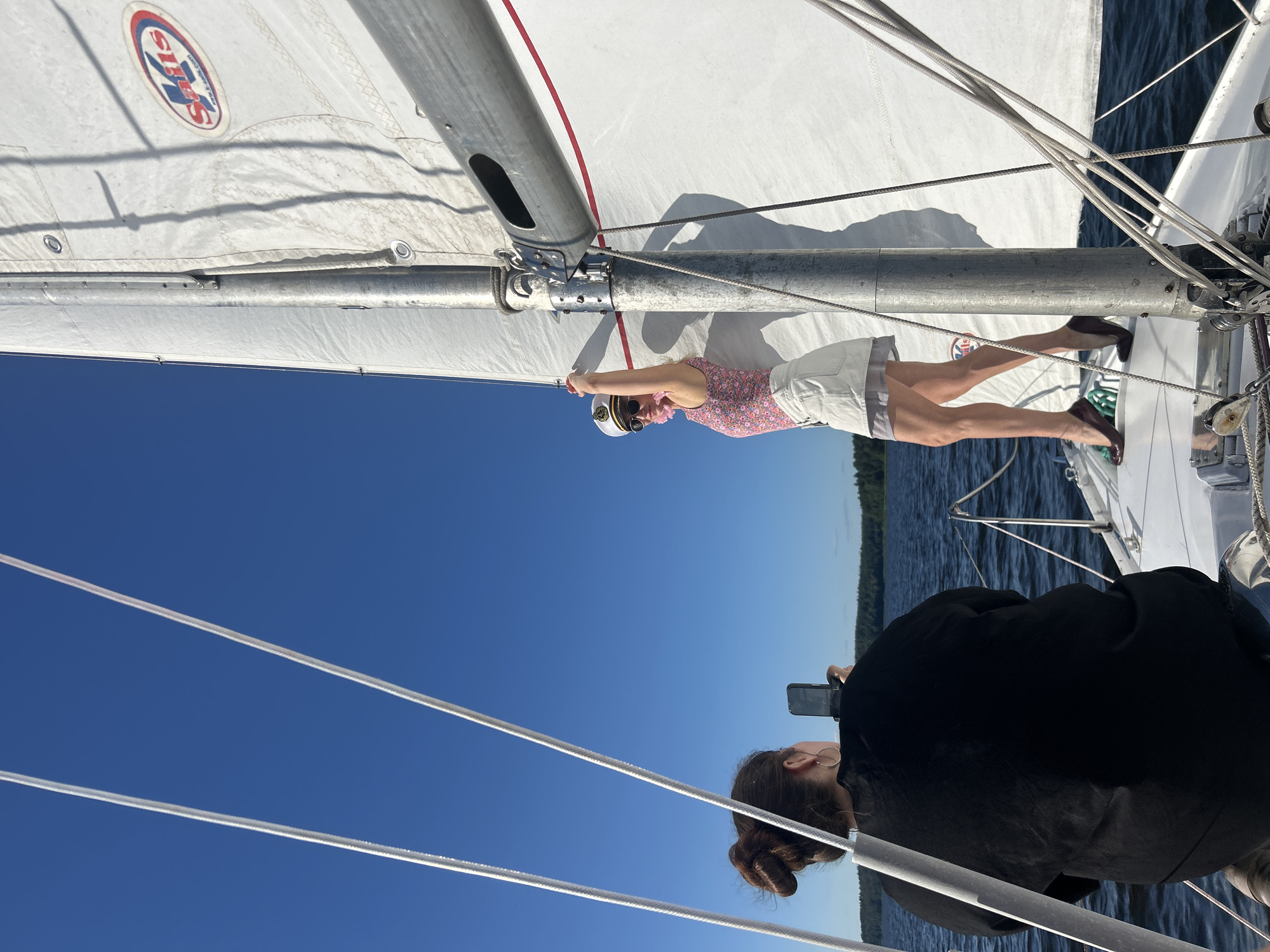 Two individuals are on a sailboat under a clear blue sky. One person, viewed from behind, wears a black top and is holding a smartphone, seemingly taking a photograph. This person has their hair styled in a bun and is wearing large hoop earrings. The other individual, positioned on the sailboat's deck, is a woman wearing a white captain's hat, sunglasses, a pink floral-patterned top, and a light-colored skirt. She is posing with her arms raised, her body angled towards the camera. The sailboat's white sail is prominent in the image, featuring a logo with red and blue circular elements and text that appears to read "K Sails". The sailboat is on a body of water, with a distant, tree-lined shore visible. The overall environment suggests a calm day on the water. The location is not Izhevsk, Russia; it is a body of water suitable for sailing.