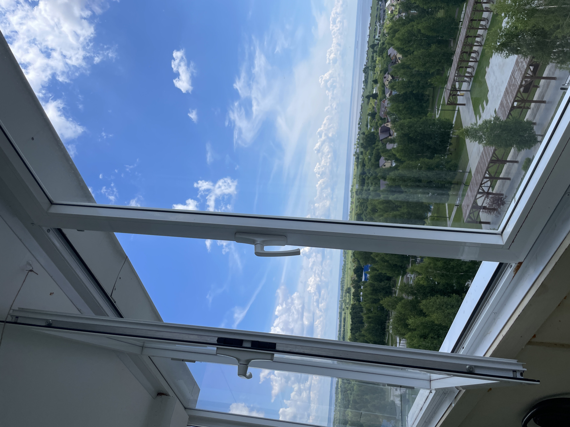 The image shows an exterior view from a window, possibly a balcony, looking out over a residential area and a body of water under a partly cloudy blue sky. In the foreground, the white frame of a window is visible, with one section slightly ajar, revealing the interior of the structure. Outside, lush green trees dominate the landscape, interspersed with houses with red and brown roofs. A paved area with wooden pergolas and benches suggests a public space or park. In the distance, a wide expanse of water stretches towards the horizon, with more land visible on the far side. The sky is a vibrant blue, dotted with scattered white clouds, some of which are wispy and streaky, indicating movement. The overall impression is one of a calm, scenic, and residential environment, likely in a suburban or rural setting on a pleasant day. The location is identified as Laishevo, Russia.