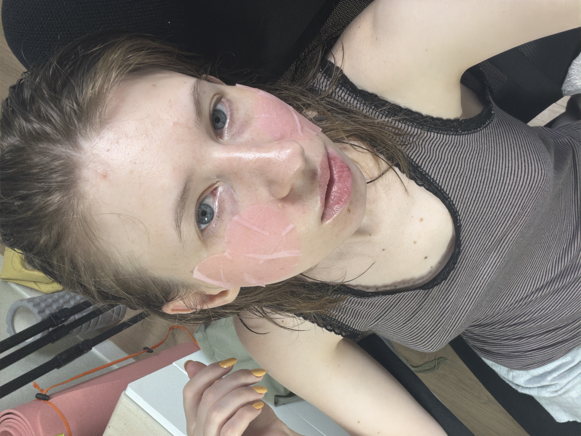 A young woman with wet brown hair and blue eyes is captured in a close-up, slightly angled selfie. She is wearing a grey and white striped tank top with black lace trim. Pink skincare patches are applied to her cheeks. Her left hand, with yellow-painted nails, is raised near her face. The background is blurred but suggests an indoor setting with yoga mats and other objects. The scene appears to be in a residential area of Dongducheon-si, South Korea.