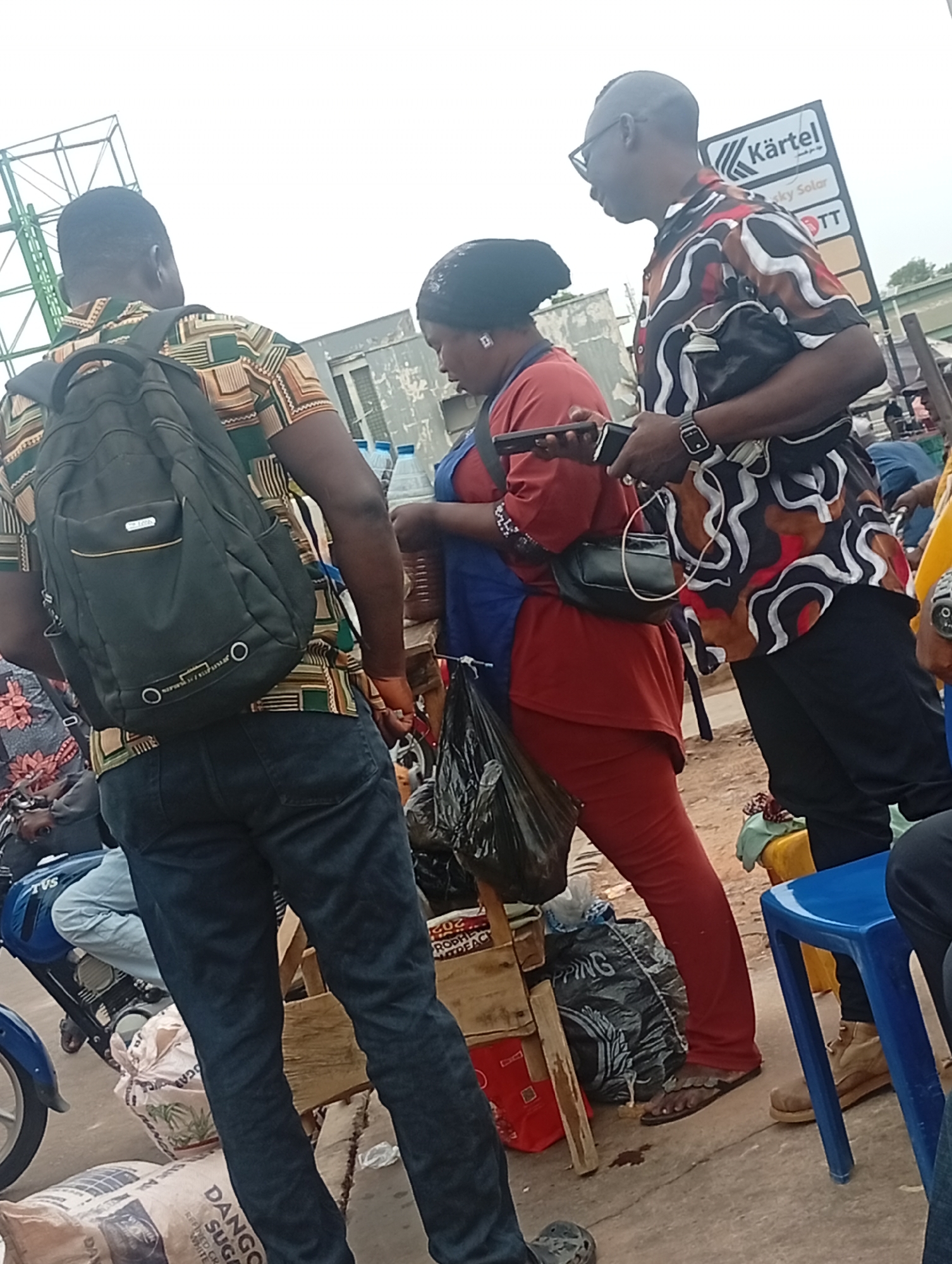 The image shows an outdoor scene in what appears to be a market area in Maiburiji, Nigeria. Three people are partially visible in the foreground. A man with a backpack and a patterned shirt is viewed from the rear. A woman in a red shirt and red pants is standing, looking at a mobile phone. A man in a patterned shirt and glasses is standing next to her, also holding a phone.
The woman has a blue apron and is standing next to a wooden stall with items for sale. Bags of "Dango Sugar" are visible on the ground near the stall. A sign with the name "Kärtel" and "Sky Solar" is visible in the background. A motorcycle is also partially visible on the left side.