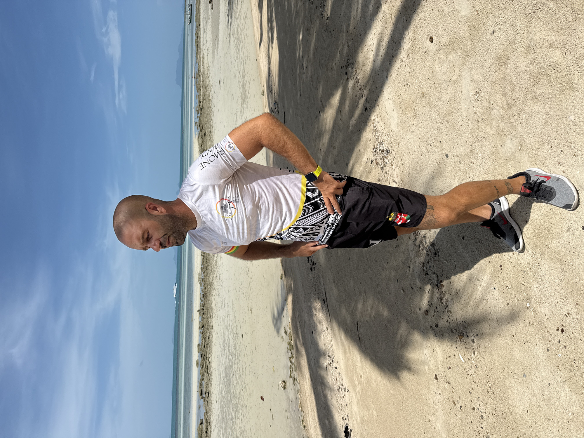 An adult male with a shaved head stands on a sandy beach in the Ko Samui District of Thailand, facing to the right and looking out at the ocean. He is wearing a white short-sleeved t-shirt with various logos and text, black athletic shorts with a patterned waistband, and gray athletic shoes with red accents. A yellow smartwatch is visible on his left wrist. The beach has sparse vegetation and scattered rocks, with palm tree shadows cast on the sand. The ocean is visible in the background, with a faint horizon line and a boat in the distance. The sky is clear and blue with scattered clouds.