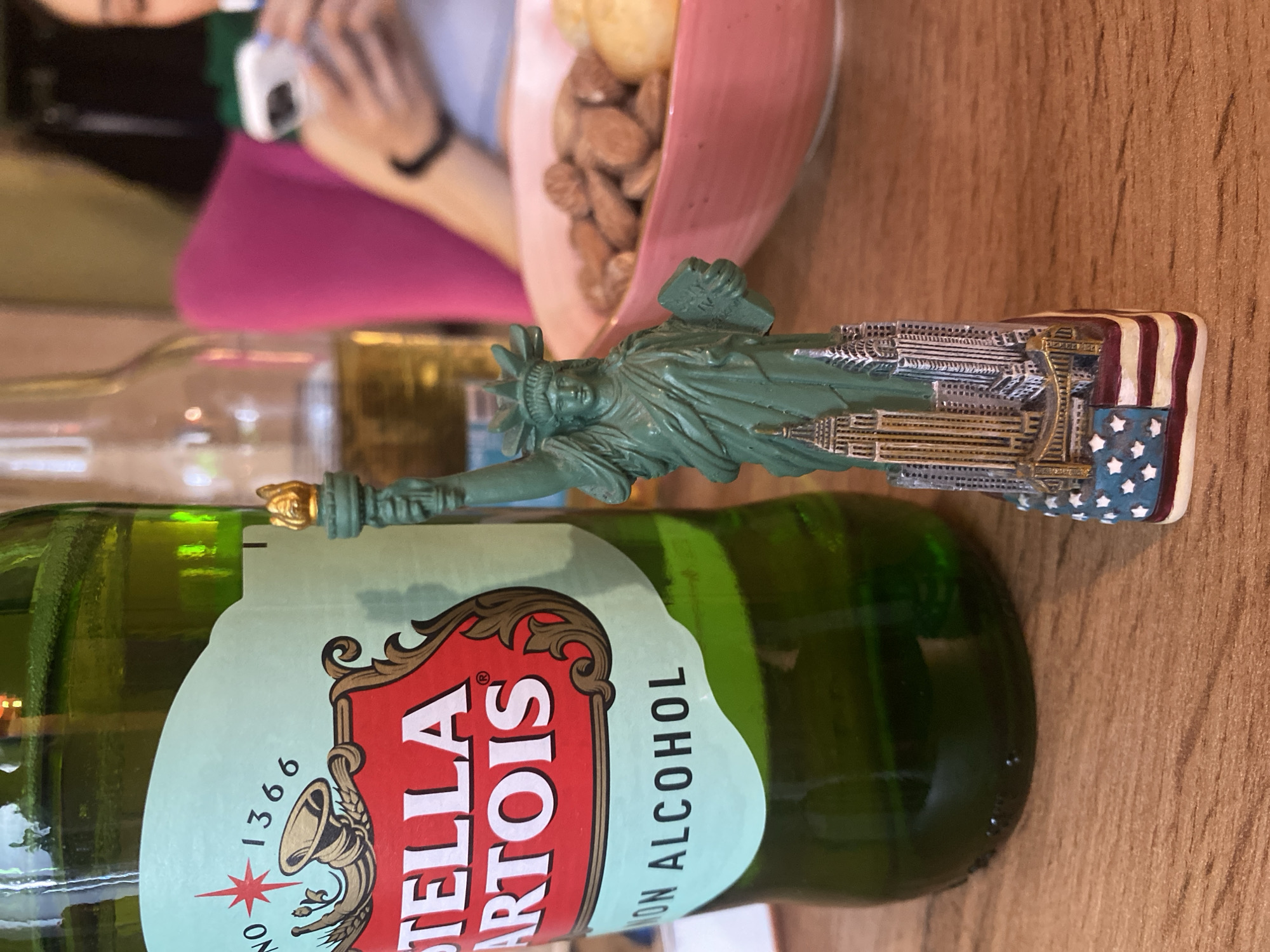 A Statue of Liberty souvenir is perched on a bottle of Stella Artois non-alcoholic beer on a wooden table. The bottle's label is partially visible. In the background, a pink bowl filled with nuts is partially in view, and a person is visible holding a cell phone. The location, Votkinsk, Russia, is not evident in the image.