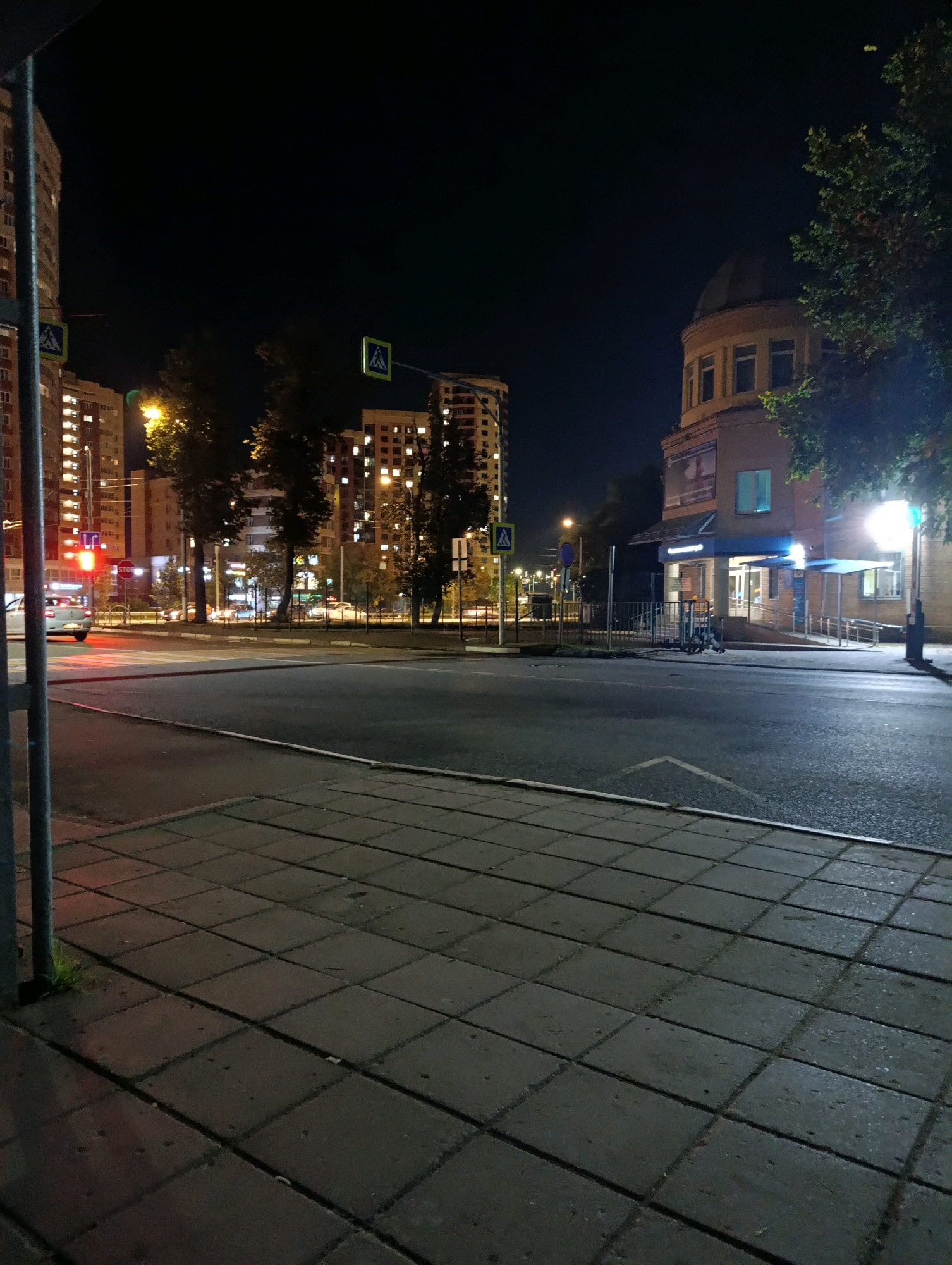 The image depicts a nighttime street scene in Solnechnogorsk, Russia. In the foreground, a gray tiled sidewalk runs along the edge of a dark asphalt road. A metal pole, likely part of a street sign or utility, stands on the sidewalk. Across the road, several multi-story apartment buildings are illuminated with many windows lit, indicating occupancy. Trees are interspersed between the buildings and along the roadside.
On the right side of the street, a circular building with a domed roof and multiple windows is visible, with a sign that appears to be for a business or institution. A streetlamp shines brightly in front of this building. Pedestrian crossing signs, featuring a blue square with a white pedestrian symbol, are present near the road. Traffic lights and streetlights are also visible, casting a yellowish glow.
A white car is seen turning at an intersection, its brake lights illuminated red. The overall atmosphere is urban and residential, with the night sky dark and clear. The sidewalk appears slightly wet, suggesting recent rain or dew.