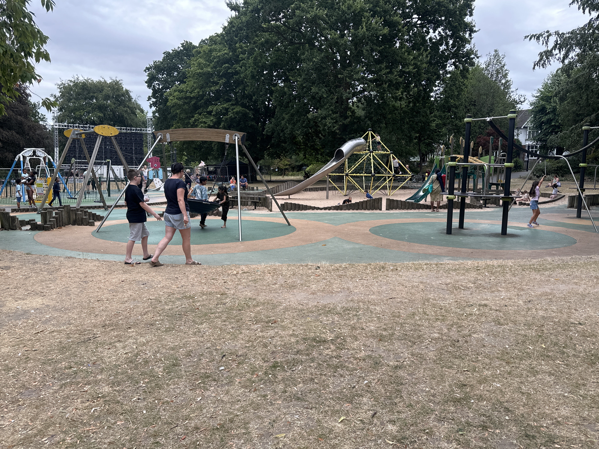 The image depicts a vibrant playground scene at a park in Rugby, United Kingdom, likely within the grounds of an event or festival, given the presence of a large stage structure with screens in the background. The playground features a variety of equipment, including swings, climbing frames, and a large metal slide connected to a yellow geometric climbing structure. Several children and adults are present, engaged in different activities. In the foreground, two individuals are walking away from the camera. One is wearing a black t-shirt and grey shorts, and the other is wearing a black t-shirt and checkered shorts. Further into the playground, a girl in a pink top and blue shorts is on a swing, and other children are seen climbing and playing on the equipment. The ground surface is a mix of sand and colored rubber matting. Surrounding the playground are mature trees and a cloudy sky, indicating an outdoor setting.