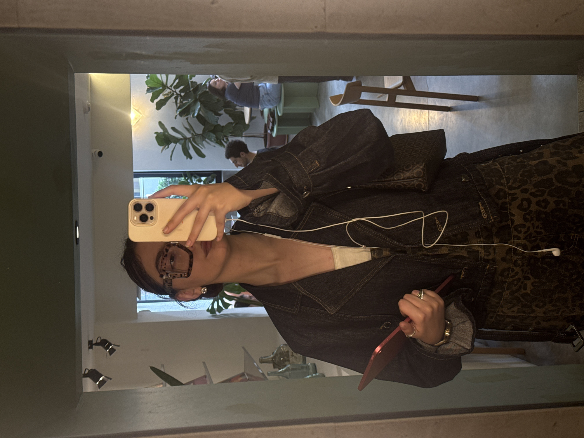 A person takes a mirror selfie. They are wearing a denim jacket over a white tank top. The jacket has leopard print details. They are wearing large square glasses, a watch, and multiple rings. They hold a red device in their right hand and a phone in their left. White earphones hang around their neck.
The mirror reflects part of a restaurant interior with a plant, other patrons, furniture, and decorative bottles on a shelf. Based on the decor, this image may be in Yerevan, Armenia.