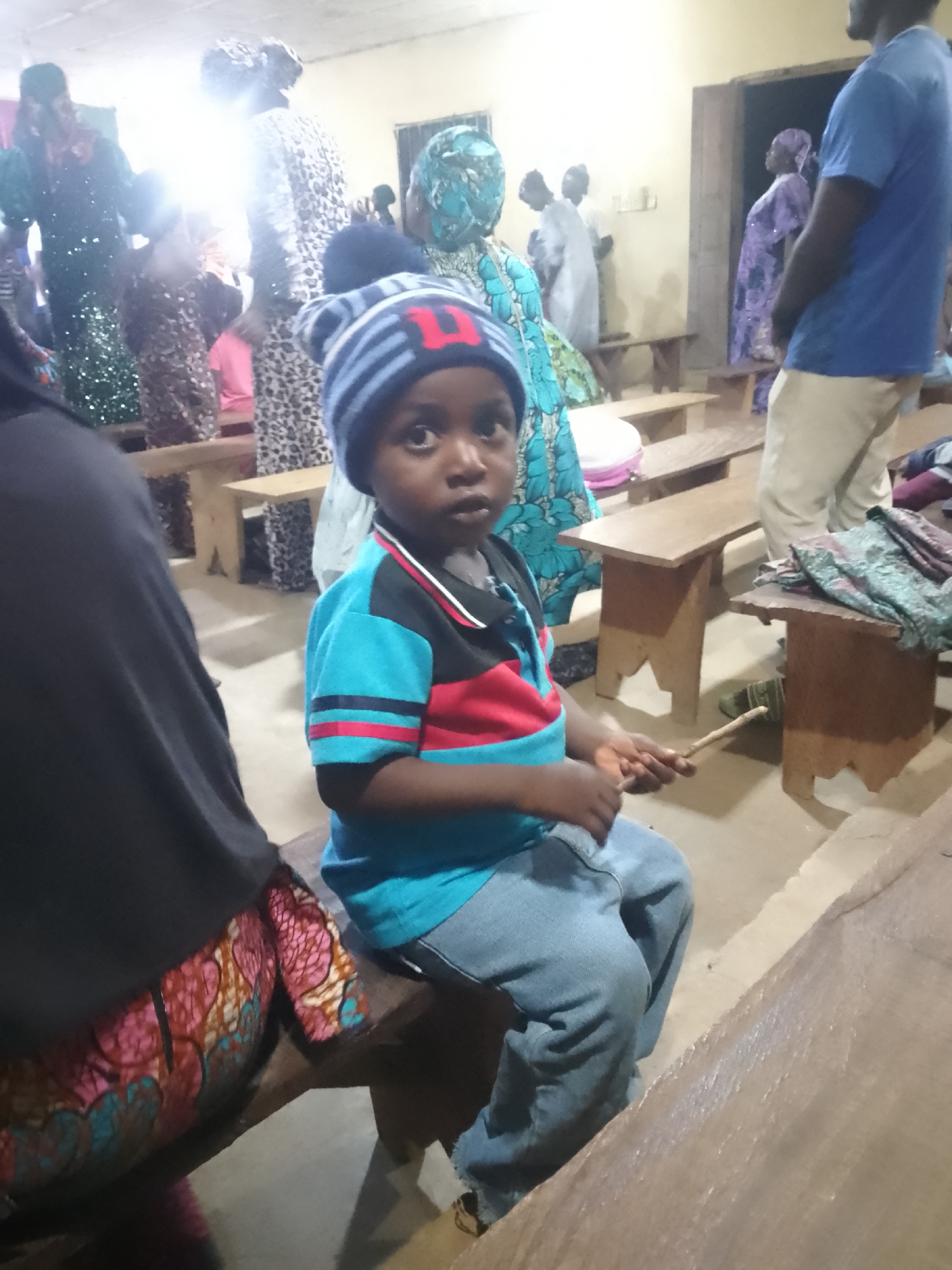 In Serti, Nigeria, a young child wearing a blue and red striped shirt and a striped beanie sits on a wooden bench. The child is holding a wooden stick and appears to be playing or tapping on the bench. The child's eyes are wide and looking directly at the camera. In the background, several adults are visible, some wearing traditional attire, suggesting a gathering or event. The environment appears to be an indoor space with simple wooden benches and walls, possibly a community hall or a place of worship. The lighting is somewhat dim, with a bright light source illuminating the background.