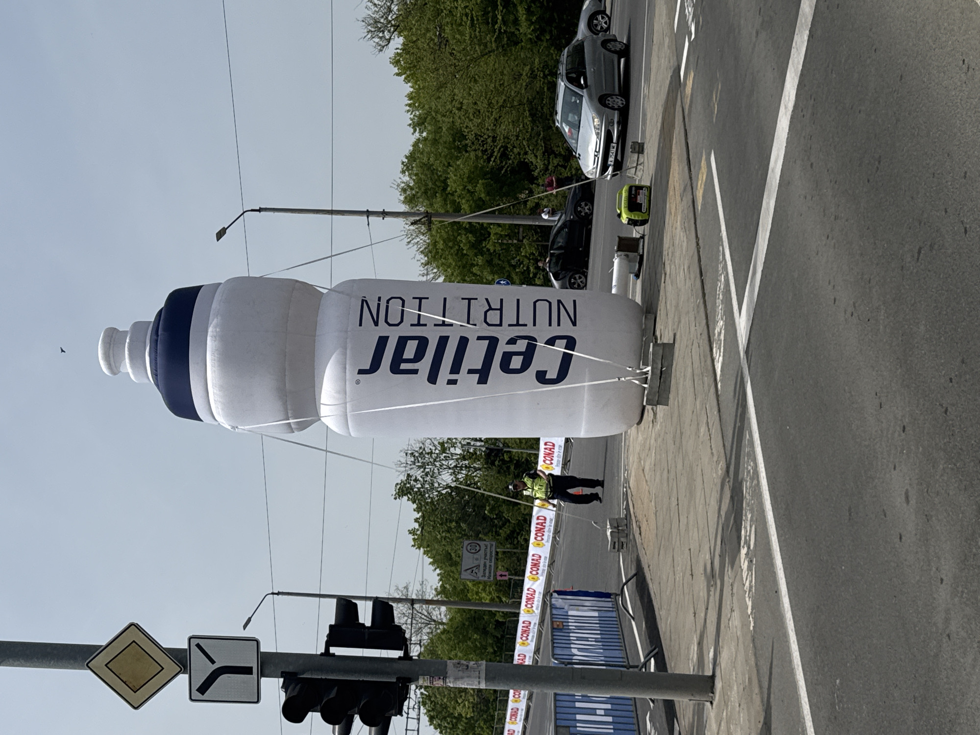 The image shows a large, inflatable bottle of "Cetilar NUTRITION" displayed in an urban environment. It's near a street with cars and road markings. There is a person standing near the bottle, appearing to manage the ropes securing it. The scene includes traffic lights, road signs, and barriers decorated with "CONAD" branding. A sign with a speed restriction symbol and Cyrillic text "Заледен участые! Намали скоростта" is present, suggesting the location is in Bulgaria. Based on the branding and signs, the location is likely in or near Burgas, Bulgaria, due to the presence of Conad grocery stores in that area and the Cyrillic text.