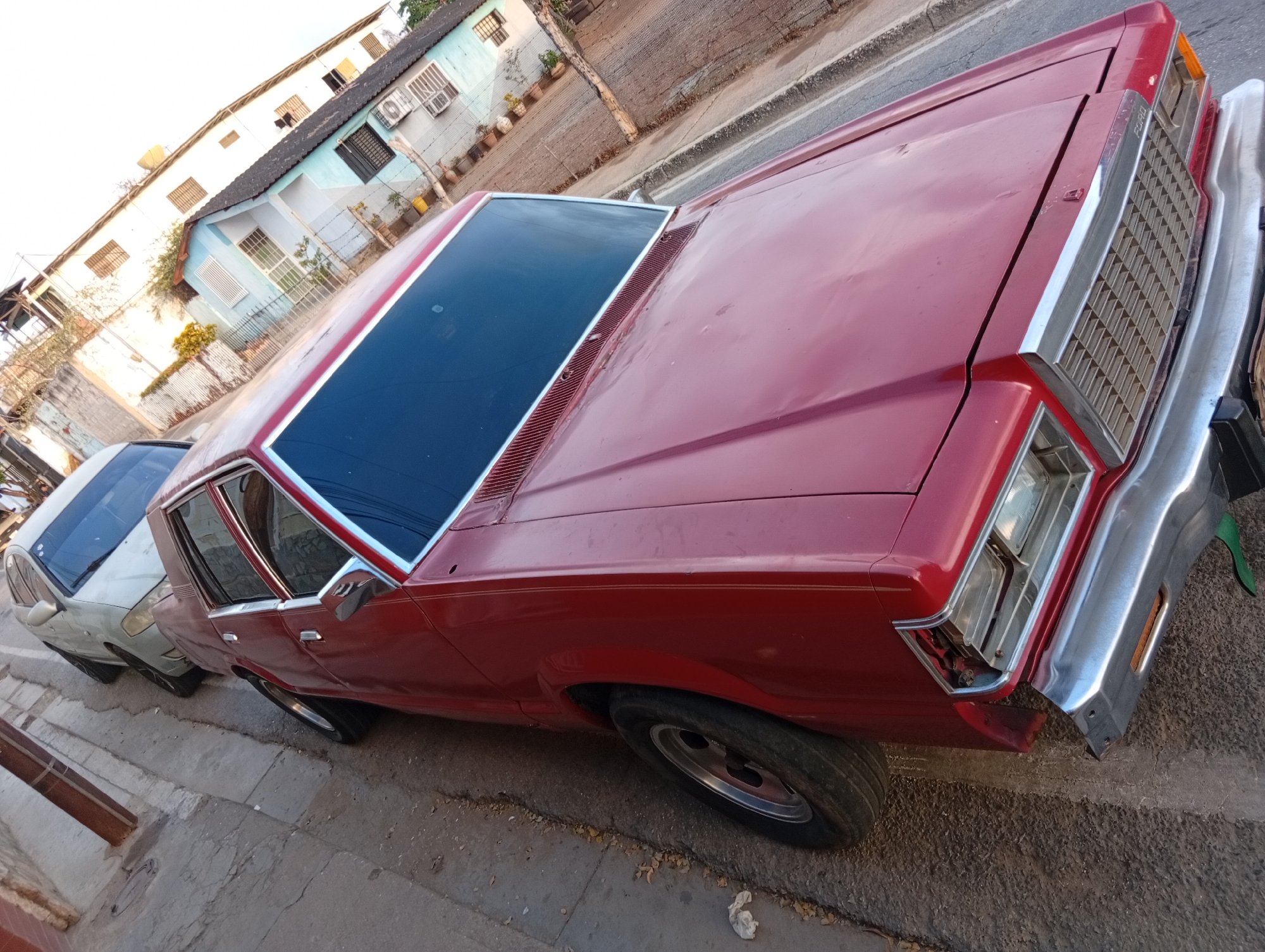 A red sedan, possibly a Ford, is parked on a street in what appears to be a residential area. The car has some visible damage to the front bumper. Another car, light-colored, is parked behind it. Buildings are visible in the background, with details such as air conditioning units and potted plants. The street appears to be paved with concrete. The location is in the city of Juan Griego, Venezuela.