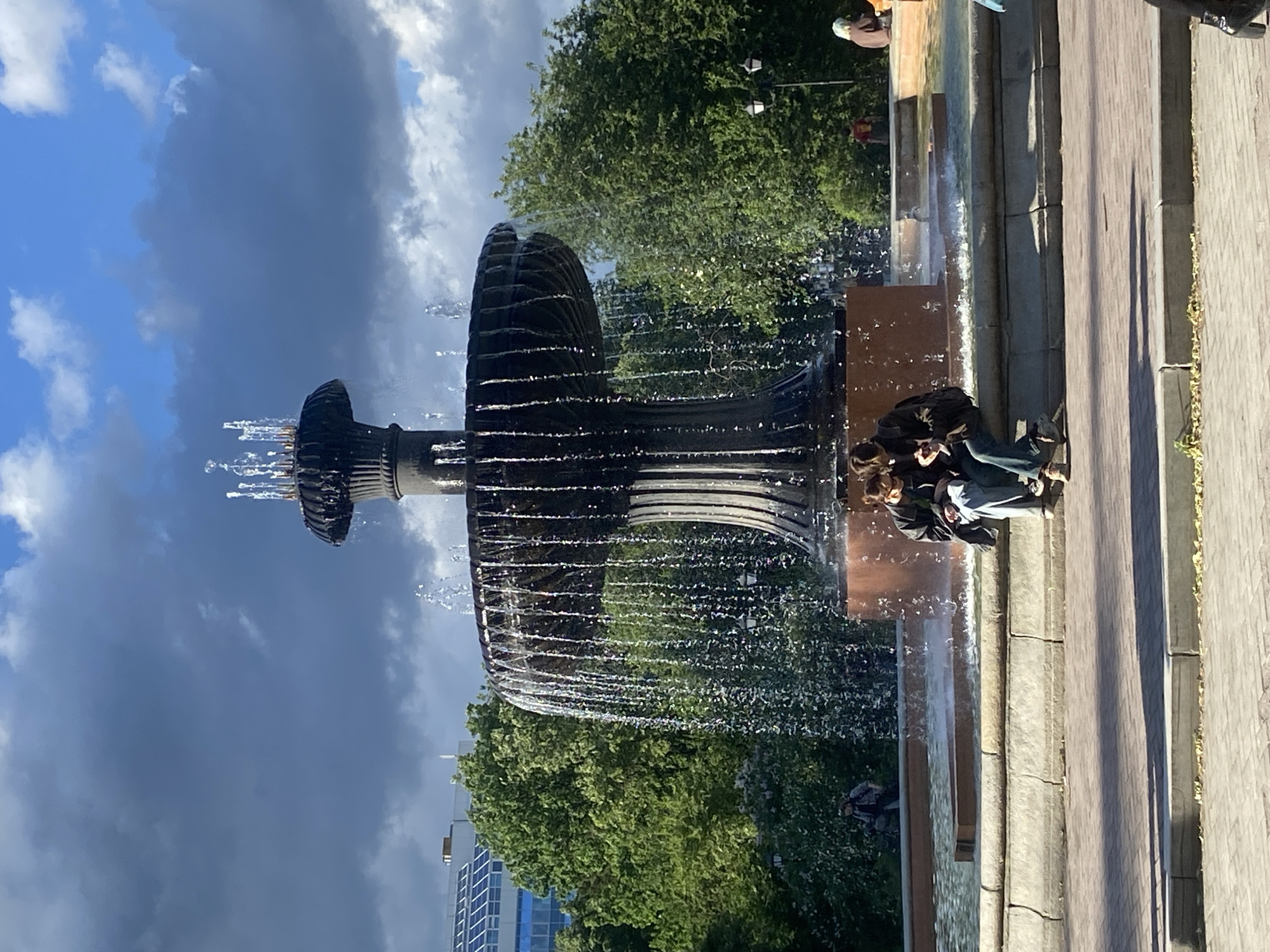 Here is a description of the image:
The image shows a fountain in a park in Yekaterinburg, Russia. The fountain has a unique design, featuring a circular structure with water cascading from it. The sky behind the fountain is partly cloudy, with blue sky and dark clouds visible. There are trees surrounding the fountain, adding to the park atmosphere. Two people are sitting near the fountain. A paved walkway is in the foreground. There are several buildings in the background.