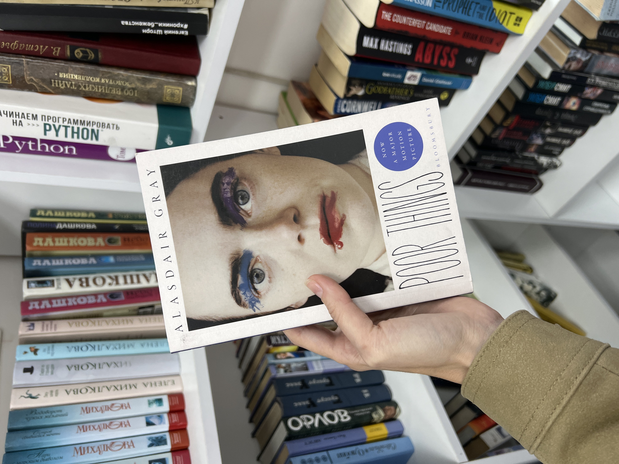 A person is holding a copy of the book "Poor Things" by Alasdair Gray in front of a bookshelf. The bookshelf is filled with books in Russian, English, and other languages. The setting appears to be a bookstore or library. The titles of some books on the shelf include "Python," "Max Hastings Abyss," "The Counterfeit Candidate," and books by Russian authors such as Dashkova and Mikhalkova. The scene is lit by natural light. The image was likely taken indoors, perhaps in a bookstore or library in Bar, Montenegro.