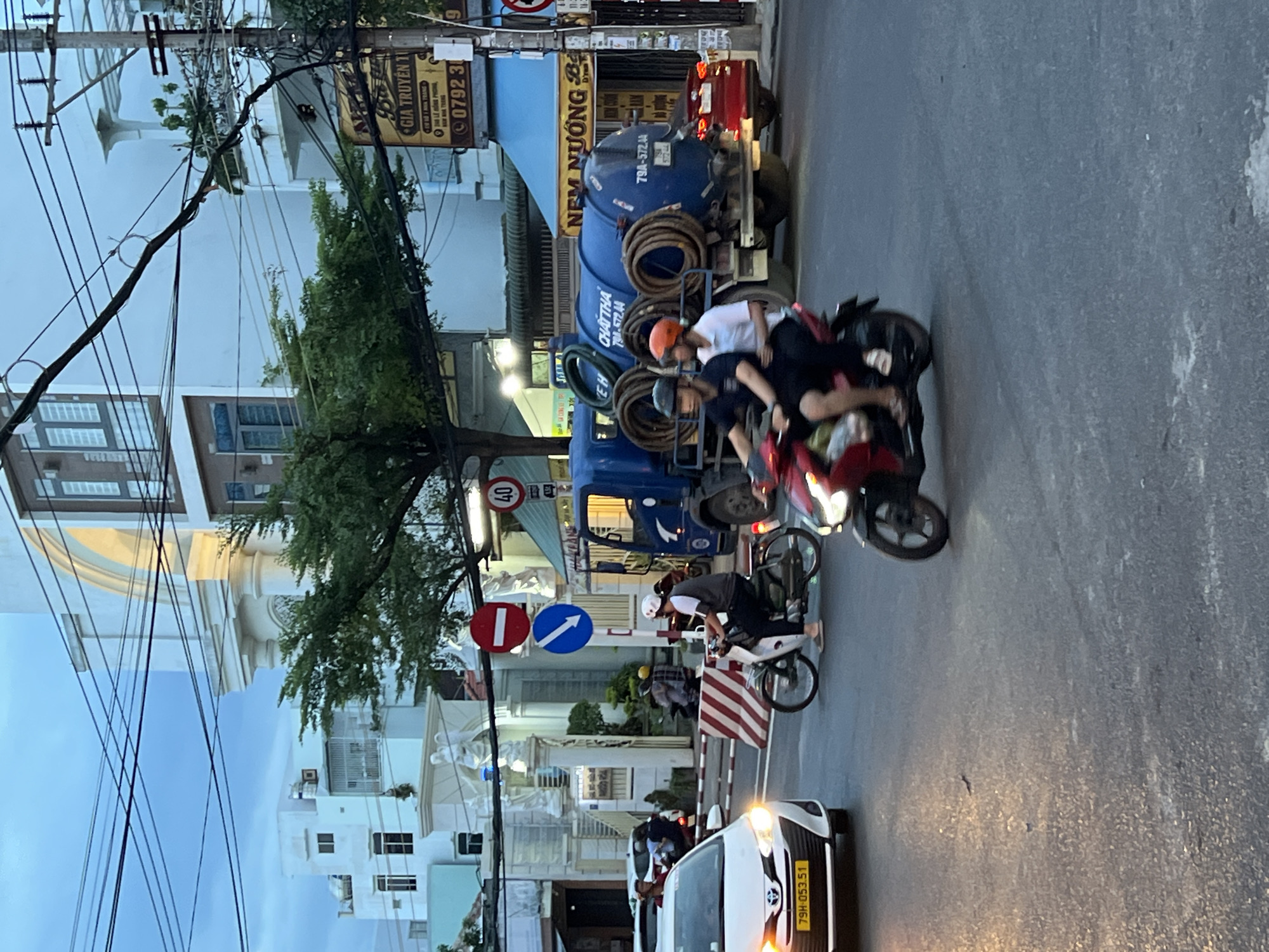 The image shows a street scene in Nha Trang, Vietnam, likely during dusk. A white Toyota sedan with license plate 79H-053.51 is in the foreground, its headlights illuminated. Two people are riding on a red motorcycle, a rider in front wearing a black shirt and helmet, and a passenger behind wearing a white shirt and orange helmet. Another person is riding a white motorcycle, wearing a grey shirt and white helmet. Behind them, a blue sewage truck with hoses is visible, and a red car is to its right. Traffic signals and street signs, including a "40" speed limit sign and a "no entry" sign, are present. Buildings with architectural details line the street. The overall environment is urban, with power lines crisscrossing the sky.
