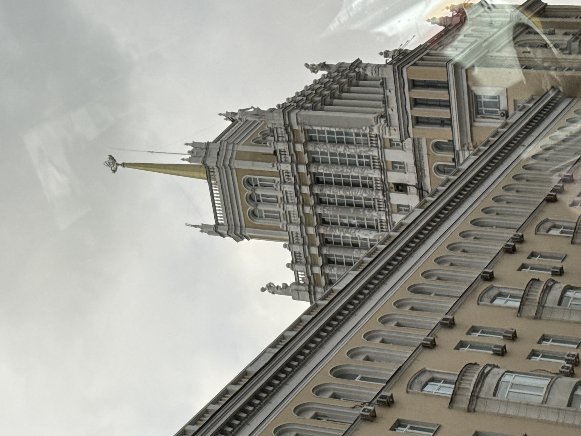 The image shows a building in Moscow, Russia, likely one of the "Seven Sisters" skyscrapers. The architectural style features a combination of Baroque and Gothic elements with classical motifs. The building is topped with a spire. The sky is overcast. The perspective is from a low angle, looking upward, which distorts the building's true proportions. The presence of reflections on a glass surface indicates the photo was taken from inside a building.
