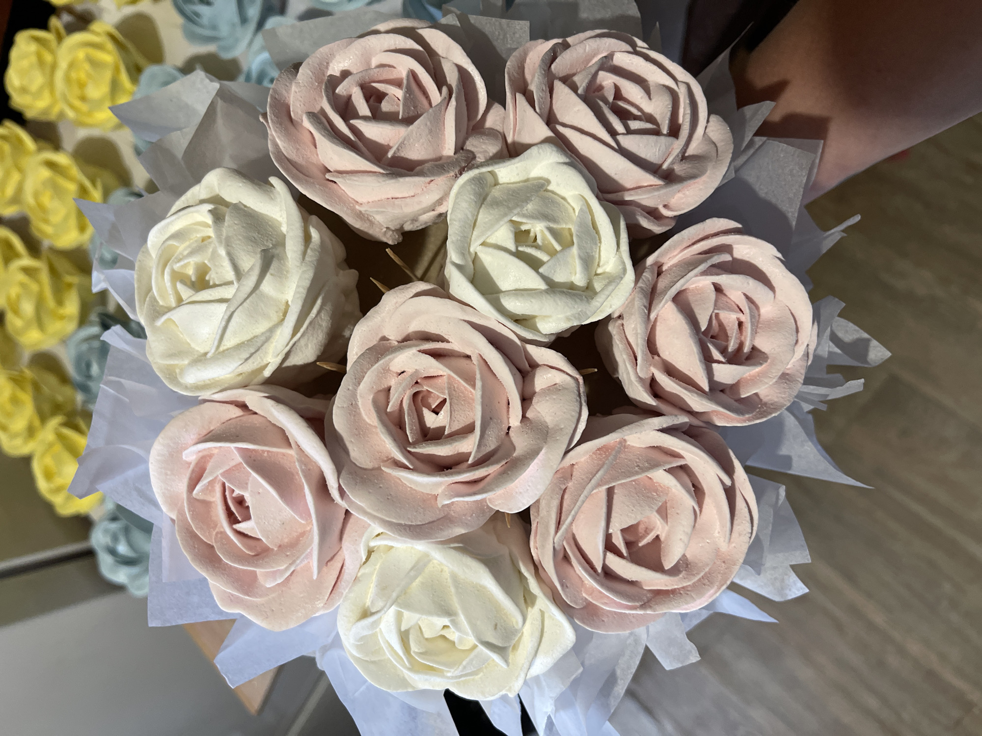A close-up overhead shot shows a bouquet of stylized roses made of confectionery, primarily pink and white, arranged on a wooden surface. The roses are crafted to mimic the petals of a rose, with some featuring a delicate pink hue and others a creamy white. They are presented in a way that suggests they are part of a larger arrangement or display, with hints of other colors like yellow and light blue roses visible in the background, further out of focus. The confectionery roses are wrapped in white tissue paper, creating a decorative effect. The lighting is bright, highlighting the textures and colors of the sweet treats. The background is blurred, suggesting an indoor setting with possibly a kitchen or display area.