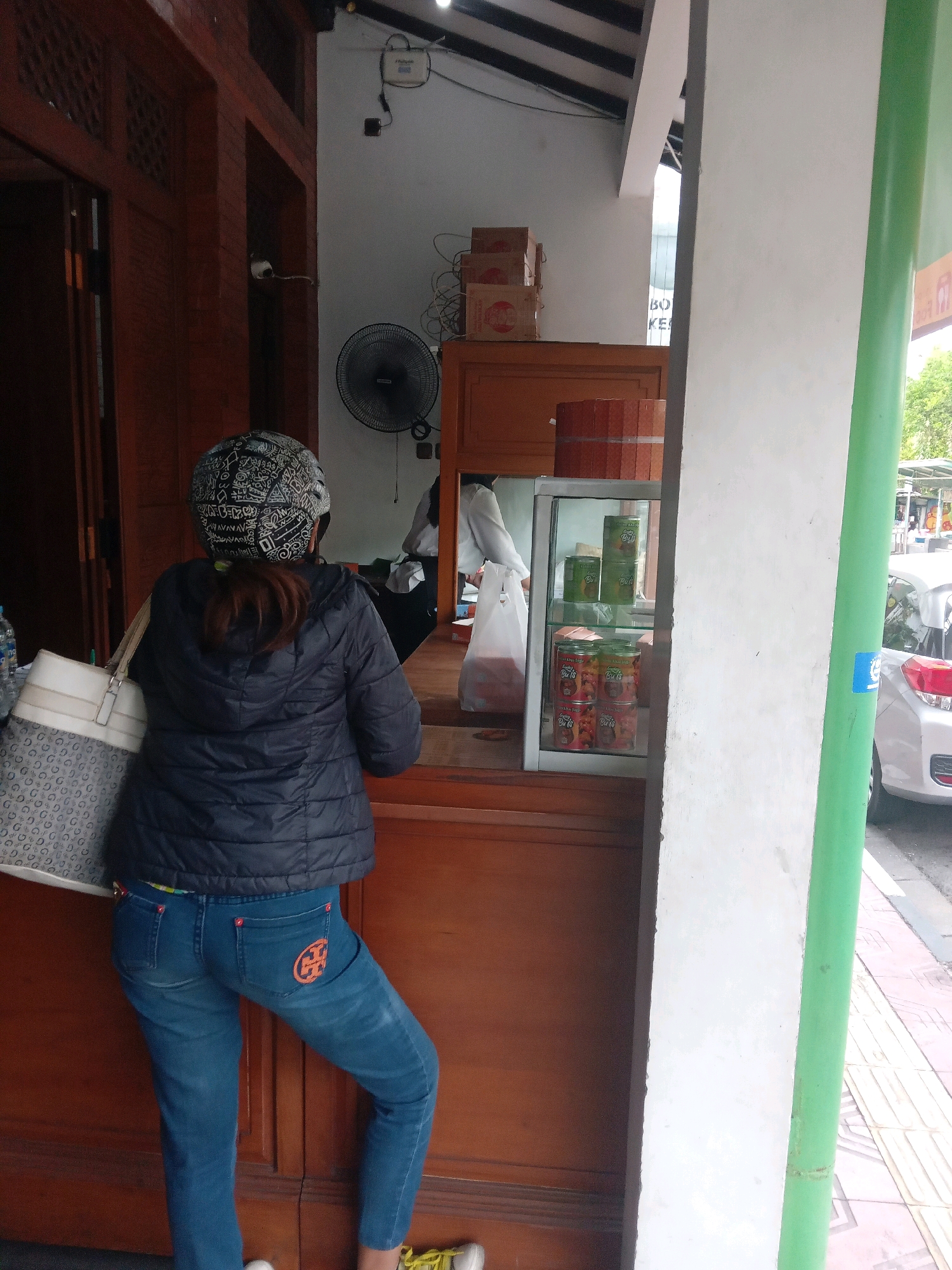 In this image, a woman wearing a black puffer jacket and blue jeans stands with her back to the camera. She has a patterned helmet on her head and is carrying a large tote bag. She appears to be waiting or browsing at a street-side kiosk. The kiosk has wooden counters and a glass display case filled with canned goods. In the background, a person in a white shirt is visible behind the counter. The scene is set in a city, likely in Yogyakarta, Indonesia, given the context. A white car is parked on the street to the right of the kiosk. The architecture suggests a warm climate with an open-air setup.