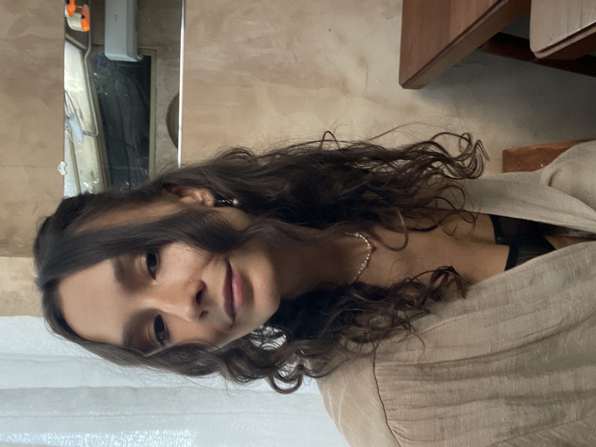 A portrait-style image, originally rotated to the left, features a young woman with long, dark, wavy hair cascading around her shoulders. She is captured in a close-up, looking directly at the viewer with a gentle, engaging smile or smirk. She wears a light beige or cream-colored, textured garment, possibly a cardigan or wrap, over a dark top. A delicate, beaded necklace is visible around her neck.
The setting appears to be indoors, likely in Mũi Né, Vietnam, during the daytime. The background includes a light-colored, possibly stucco or textured wall. To her left, a window or mirror reflects an outdoor scene showing dark foliage. Above the reflection, a white rectangular object, resembling an air conditioning unit or light fixture, is mounted on the wall. Wooden elements, possibly shelving or a frame, are visible in the upper right of the frame. A sheer white curtain can be seen in the lower left corner. The lighting is soft and natural, illuminating her face. There is no visible text in the image.