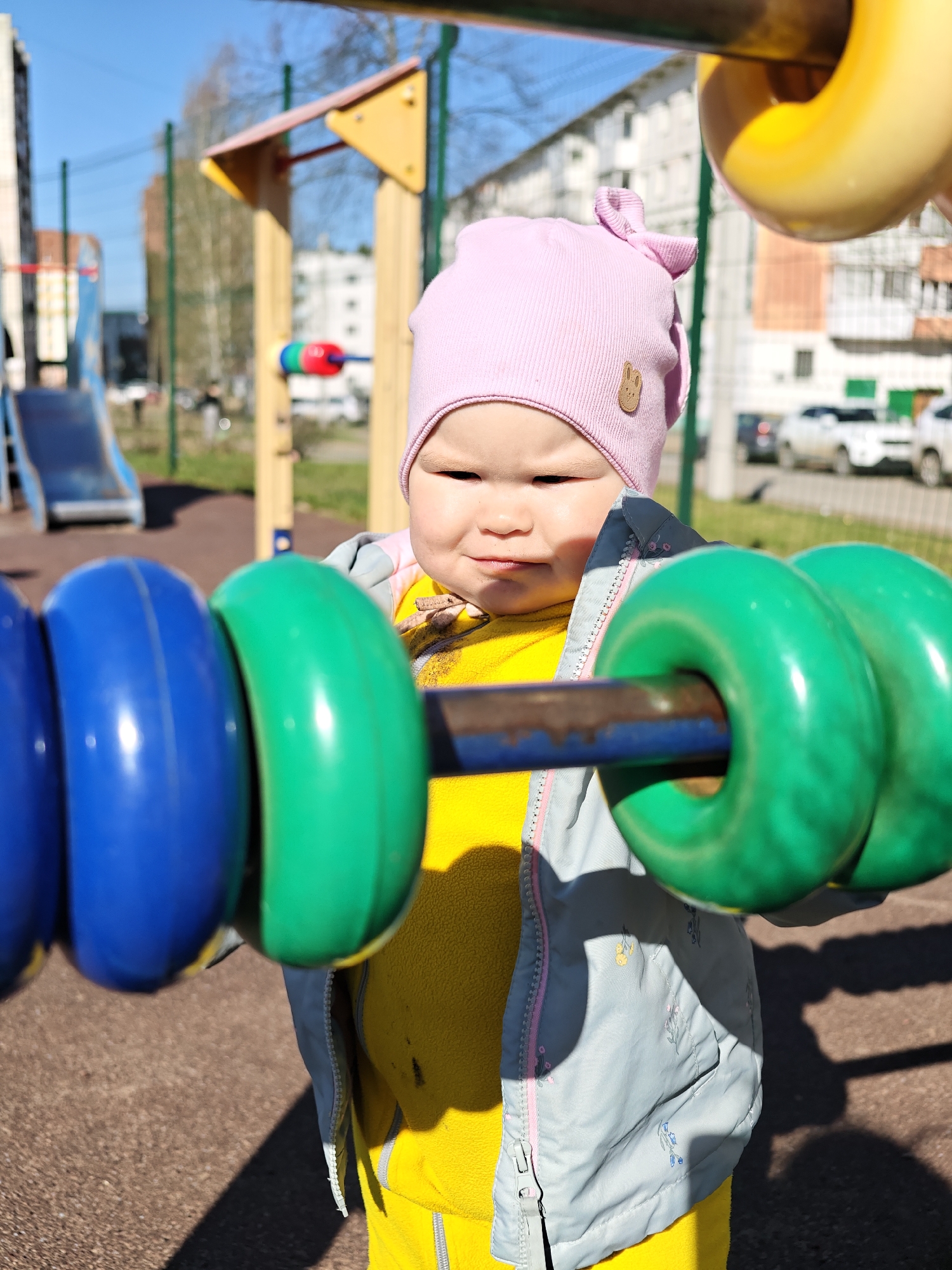A young child in Chaikovsky, Russia stands on a playground facing the camera. The child is wearing a pink hat with a small bunny logo, a yellow fleece jacket, and a light blue jacket. The child is standing near a play structure with large, colorful rings. Blue rings are on the left and green rings are on the right. Yellow rings are visible in the upper right corner. In the background, there's a slide and an apartment building. The ground is brown, likely a rubberized safety surface. The lighting suggests it's a sunny day.