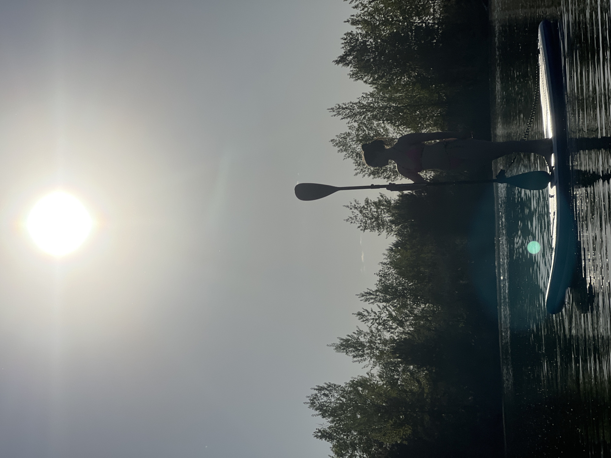 A silhouetted figure stands on a paddleboard, utilizing a paddle in the water. The trees on the bank are dark green, while a bright sun dominates the left side of the frame, obscuring details. The water reflects the sun's glare. The location is implied to be near Novyi, Russia, based on the user prompt.