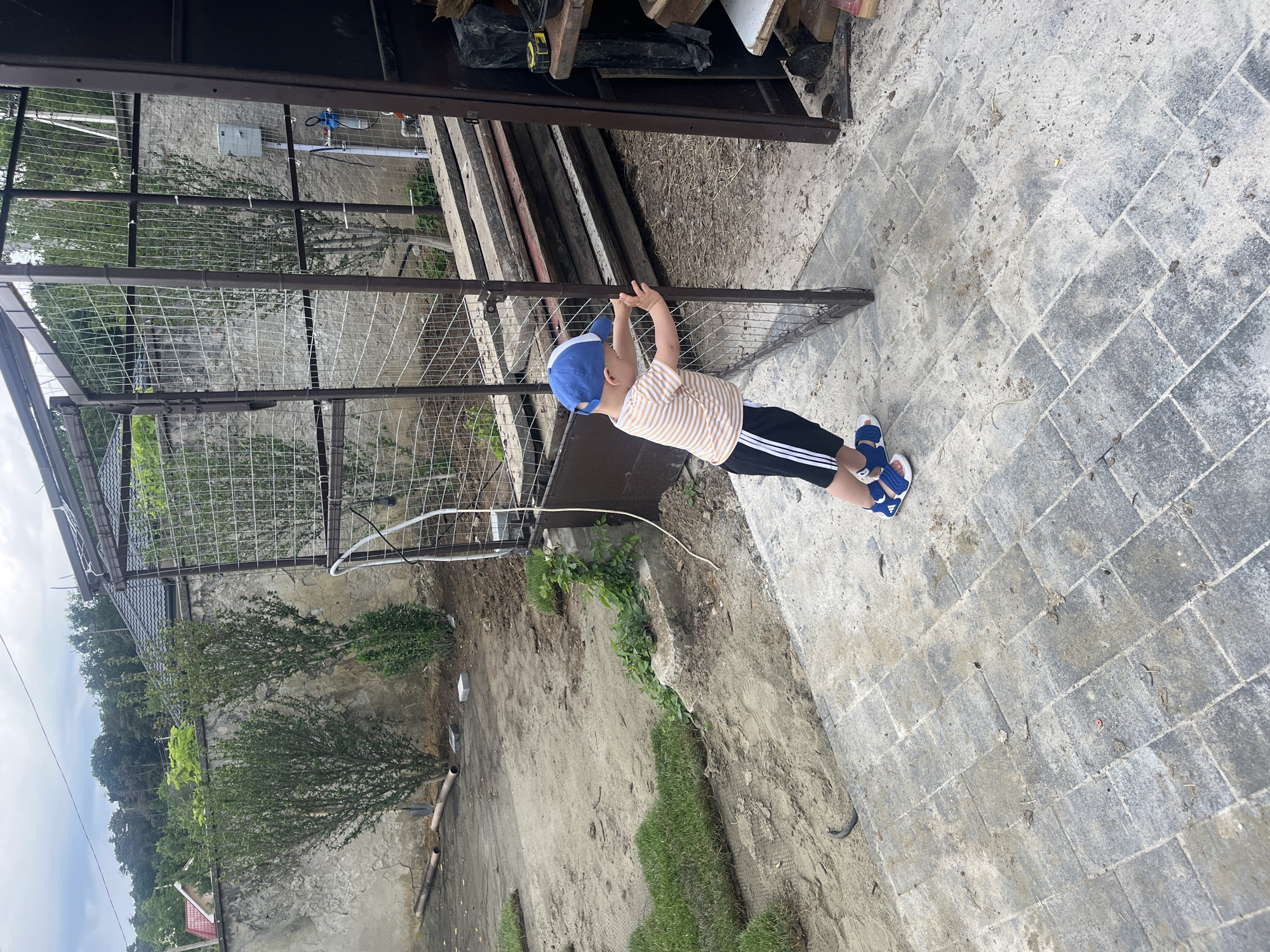 A young boy in a striped shirt, black shorts with white stripes, a blue and white baseball cap, and blue sandals leans against a partially opened metal gate. The gate is part of a cage-like structure made of metal posts and wire mesh, with a dark brown metal door on the right. The boy is positioned on a paved area made of grey rectangular stones, which is dirty and scattered with debris.
Behind the gate, to the left, is a weathered concrete wall with two neatly trimmed green bushes in front of it. There are also two pieces of brown pipe and a shovel on the ground near the wall. Further left, a section of lawn is visible, with rolled sod placed on the dirt. In the background, trees and houses with tiled roofs are visible under a cloudy sky. To the right of the gate, a dark brown cabinet or structure is visible, with shelves containing wood planks, a tape measure, and other items. The overall environment appears to be an outdoor yard undergoing landscaping or construction. The location is identified as Onokivtsi, Ukraine.