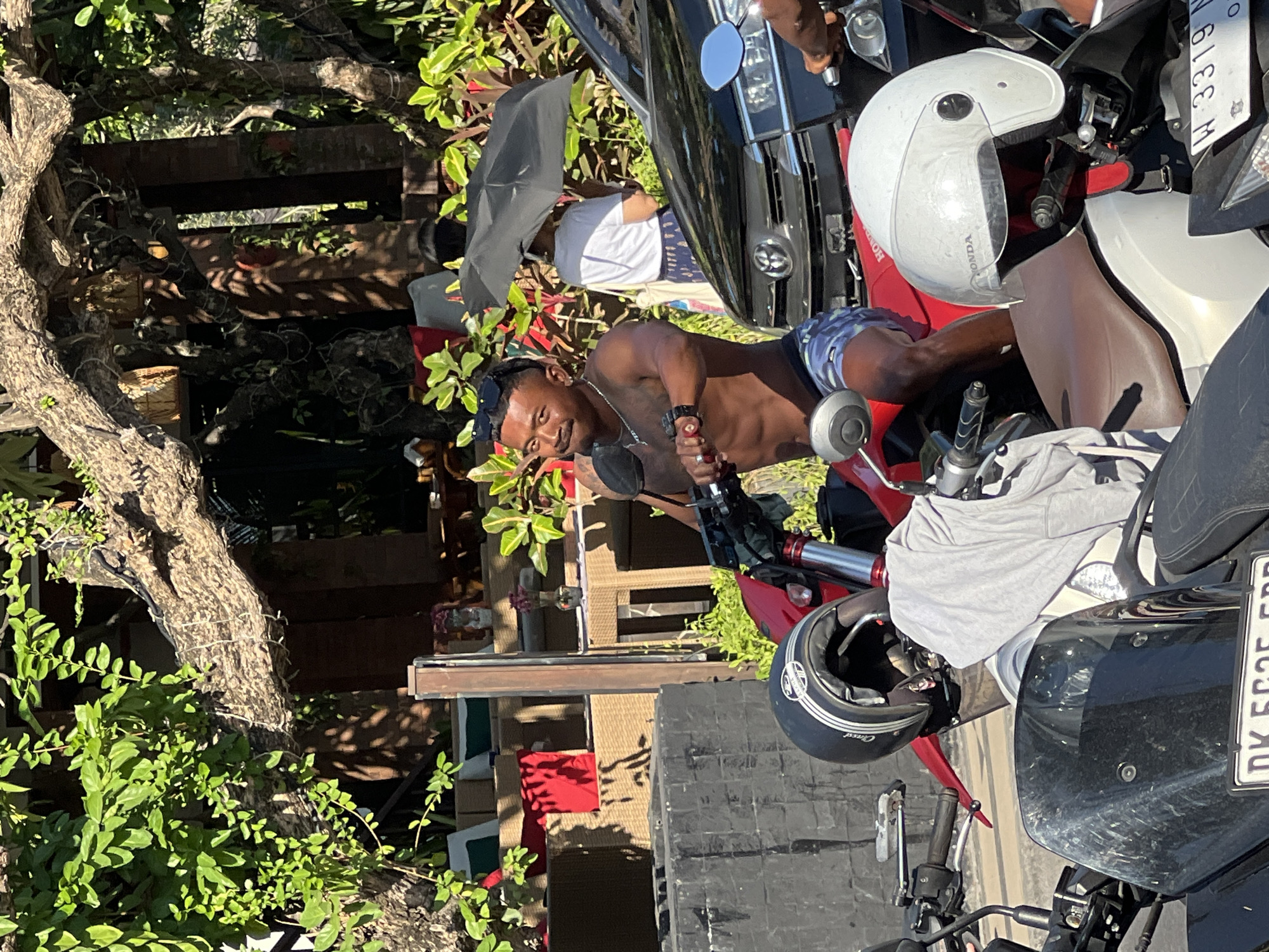 A shirtless man with tattoos, wearing blue and white patterned shorts and sunglasses, sits on a red motorcycle. He is looking at the camera with a slight smile. In the background, a person holding a black umbrella stands near a parked black car. The scene appears to be a street in Kuta, Indonesia, with lush green foliage and a rustic wooden structure. Several other motorcycles are parked nearby, some with helmets on them. The sunlight suggests it is daytime.