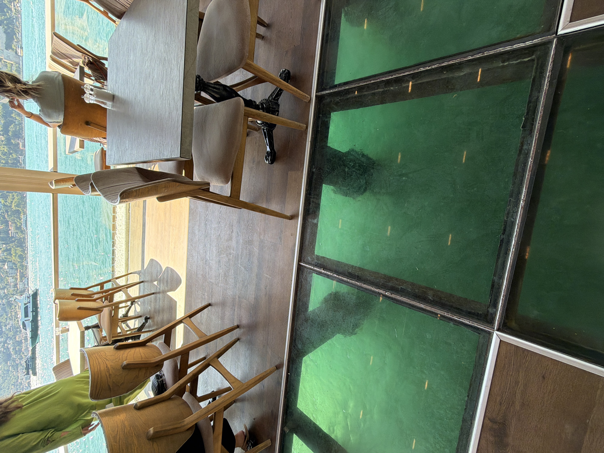 The image depicts a well-lit, modern restaurant interior with a prominent glass floor revealing a green-tinted body of water below. The restaurant appears to be situated in Beykoz, Turkey, overlooking the Bosphorus Strait, with boats visible in the distance.
Inside the restaurant, several wooden chairs with beige cushioned seats are arranged around tables. One table is covered with a textured grey surface. In the foreground, a person wearing a green garment is partially visible, standing near a chair. Further into the restaurant, another person, viewed from the back, is seated at a table, facing the expansive window and the water view. Two salt and pepper shakers are placed on a table.
The glass floor is divided into rectangular sections by metal framing. Beneath the glass, the water has a vibrant green hue, and shadows of people are visible, suggesting they are walking on the glass floor above. The overall ambiance is one of contemporary dining with a unique architectural feature.