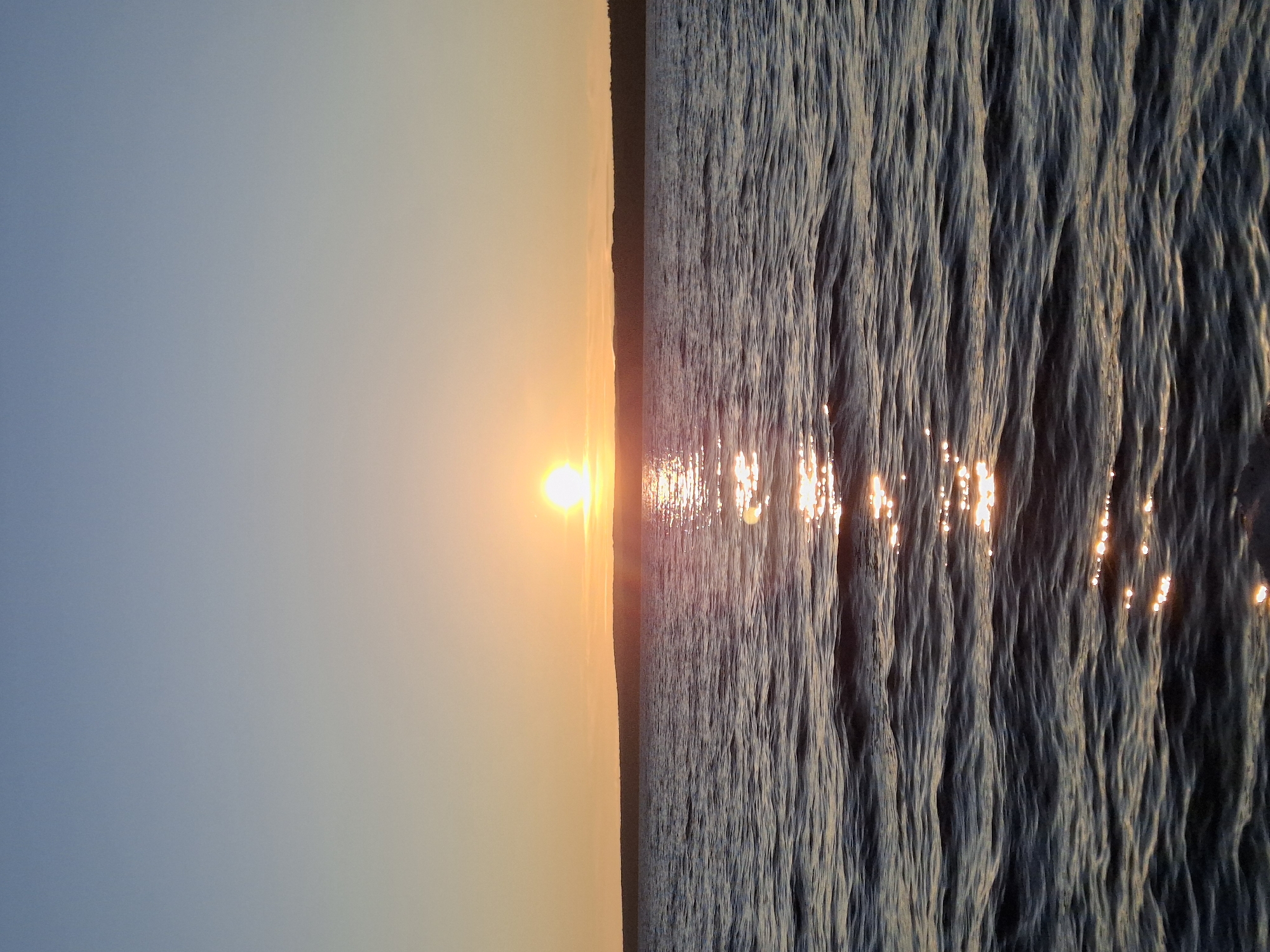 The image depicts a sunset over a body of water, likely a river or lake, with a faint tree line visible on the horizon. The sky transitions from a pale blue at the top to a warm orange hue near the horizon, where the sun is setting. The water's surface is rippled, and the setting sun casts a shimmering reflection across it. The scene appears tranquil and natural, with no visible people or man-made structures in the immediate vicinity. Given the context of Chaikovsky, Russia, this scene could be set on the Kama River, which flows through the city and is a prominent natural feature. The overall impression is one of natural beauty at dusk.