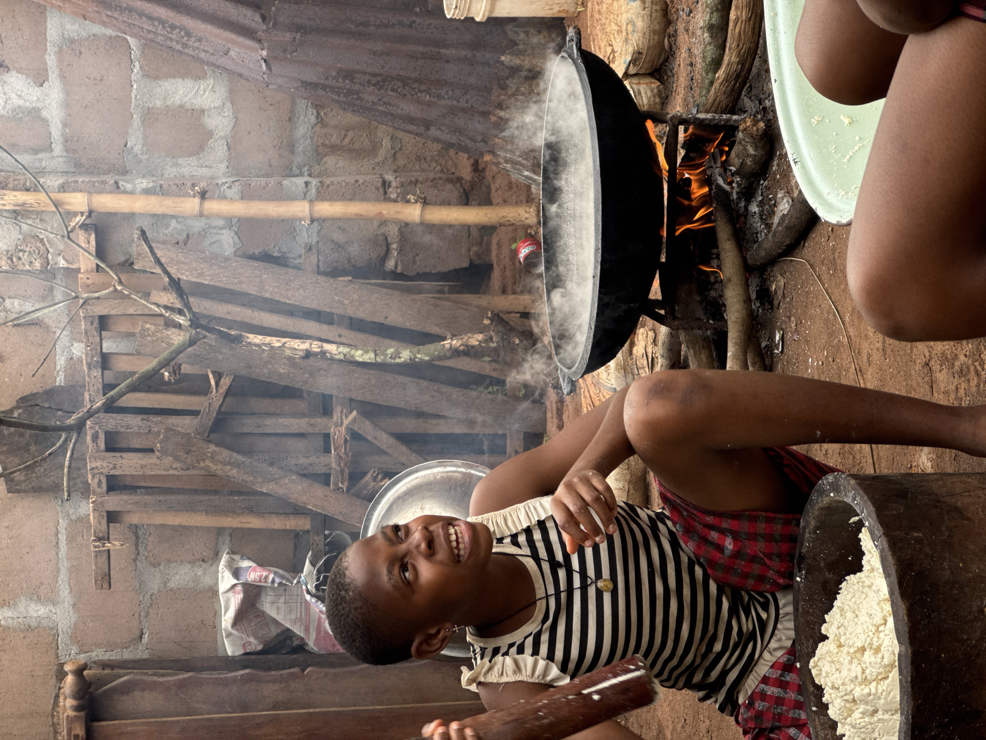 This media file captures a vibrant scene of food preparation in an outdoor or semi-outdoor kitchen setting in Eziudo, Nigeria. The image is composed from a slightly elevated perspective, looking down into the activity.
The central subject is a young person, likely female, with short hair, wearing a striped short-sleeved top with ruffled shoulders and a red and black plaid skirt. She is smiling broadly and looking towards the camera, holding a wooden pestle. Her hands and face show some traces of the white substance being processed.
To her lower right, a large, dark wooden mortar is visible, filled with a white, granular or pasty substance, likely processed cassava or a similar food staple.
In the background, a large, black metal pot is set over an open wood fire, with visible flames and rising steam, indicating active cooking. Beneath the pot, a red "Sprite" can is partially visible. Further to the right, the lower legs of another person are visible next to a light green basin, suggesting multiple individuals are involved in the activity.
The setting features a rustic environment with a wall constructed from concrete blocks and cement rendering. Wooden planks and branches form part of a lean-to structure or shelving in the background, along with a bamboo pole spanning horizontally. A white sack with "2.5M" and other text is also visible on a shelf. The ground is a dirt surface, characteristic of an outdoor workspace. The natural lighting suggests it is daytime. The overall scene conveys a sense of communal activity and traditional food preparation.