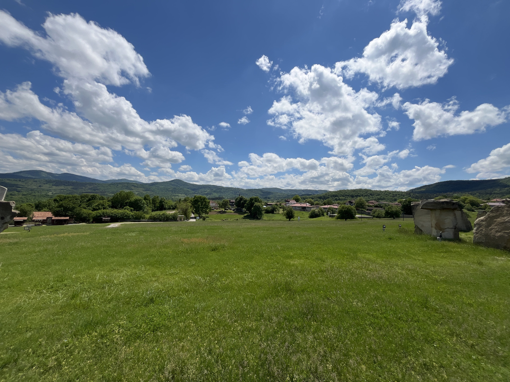 The image shows a panoramic view of the Stone Mushrooms landmark in Gardevtsi, Bulgaria. A wide expanse of green grass fills the foreground, transitioning into a distant village nestled amongst trees at the base of rolling, forested hills. The sky above is bright blue and dotted with puffy, white cumulus clouds. To the right of the frame are some natural rock formations called stone mushrooms. Some houses and trees are on the left.