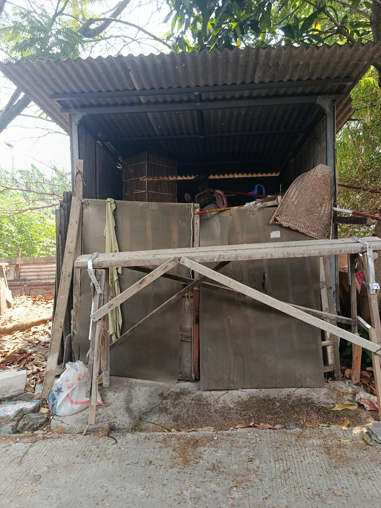 The image depicts an outdoor scene in Wanasari, Indonesia. In the center, there is a makeshift, open-sided shelter with a corrugated metal roof supported by metal beams. The walls appear to be made of a dark, opaque material, possibly sheet metal or thick plastic. Inside the shelter, a birdcage is visible, along with various stored items including what looks like a woven mat or basket, a blue object, and other unidentifiable items. In front of the shelter, there is a makeshift wooden scaffolding structure constructed with rough-hewn lumber. A light-colored plastic bag with red and blue markings lies on the ground near the base of the shelter. The ground in front of and around the shelter is concrete with debris, including fallen leaves and small branches. The area is overgrown with foliage, with trees and branches visible above and to the sides of the shelter, suggesting a somewhat neglected or rural setting within the city.