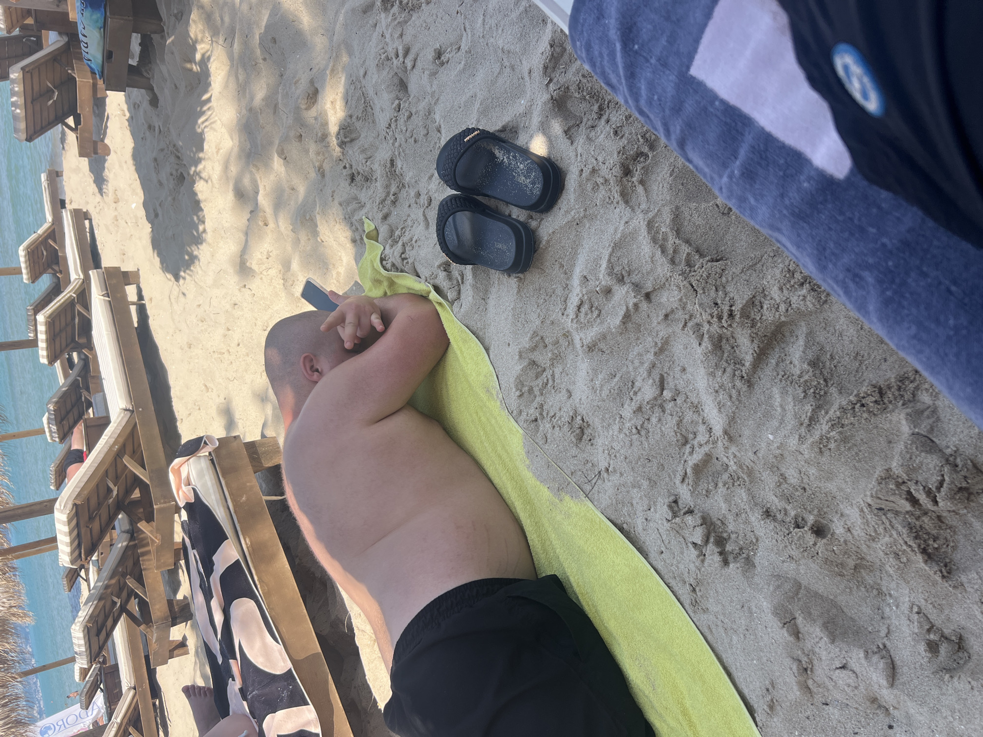 A high-angle photograph captures a sandy beach scene in Kranevo, Bulgaria, under bright sunlight. The immediate foreground features the lower legs and lap of a person, clothed in dark blue shorts or trousers that display a white rectangular patch and a partially visible blue and white circular logo on the fabric. This person is positioned as the photographer.
To the left of the frame, a bald, shirtless individual is lying prone on a bright yellow rectangular towel placed on the light-colored sand. This person is wearing black swim shorts. Their head is turned slightly to the left, and they are holding a dark-colored smartphone with both hands, with their forearms resting on the towel and sand. The skin on their visible back and left side displays faint, reddish, irregular marks.
A pair of black, textured slides, each with a subtle gold-colored accent, are resting on the sand near the lying individual's head, partially covered in sand particles. The sand in the mid-ground shows signs of disturbance, including footprints, and is marked by dappled shadows cast from overhead foliage or structures.
In the background, multiple wooden beach loungers with light-colored, possibly striped, cushions are arranged. Several straw-thatched umbrellas provide shade over these loungers. At least two additional individuals are visible: one on the far left with only their legs discernible on a lounger, and another on the far right, wearing red swim shorts and facing away from the camera towards the sea.
The far background reveals calm, blue-green ocean water extending to a distinct horizon line under a clear, bright sky. A small, indistinct human figure is present in the water. On the far left, a white inflatable or banner with dark Cyrillic lettering is partially visible behind a lounger.