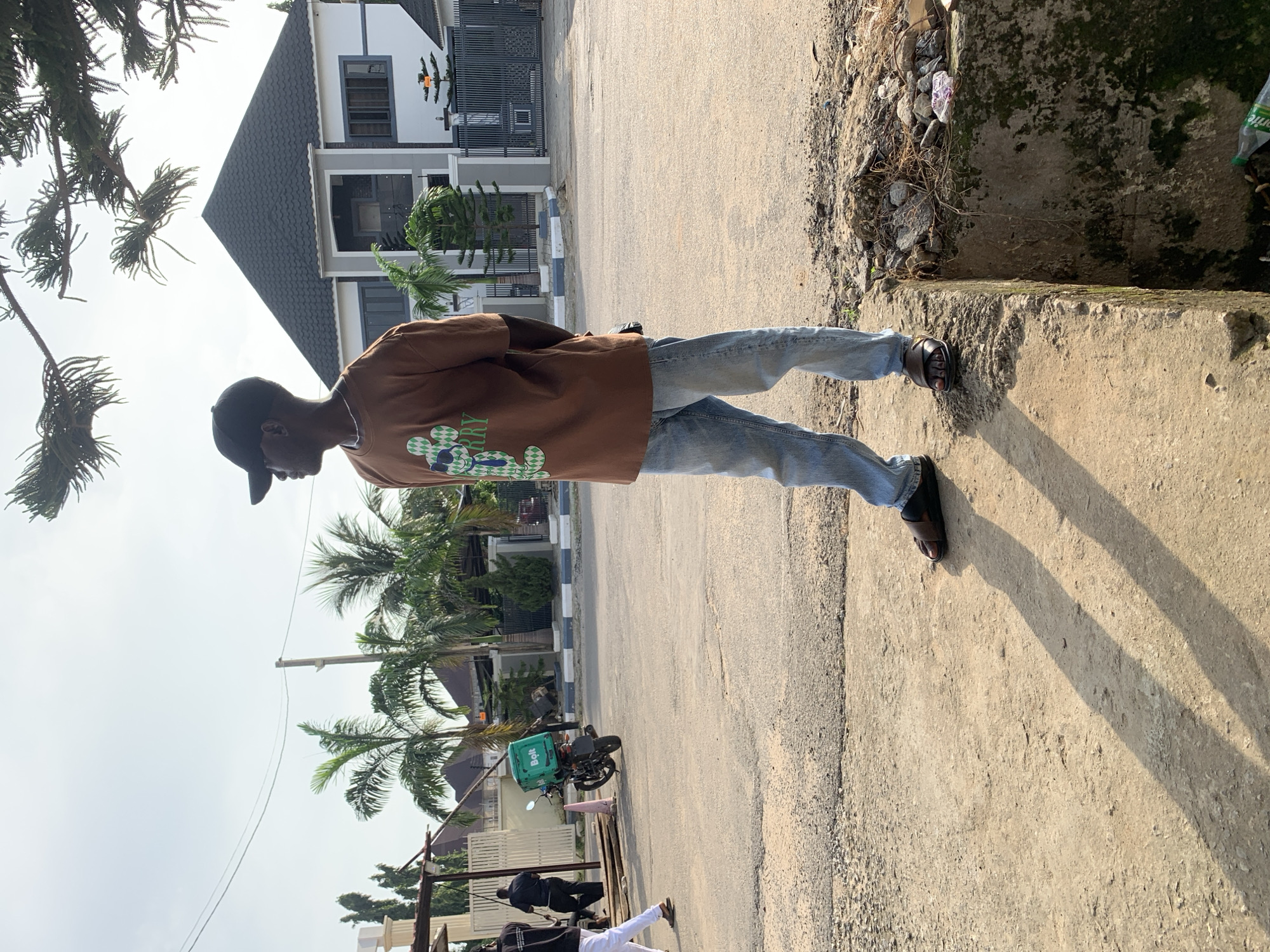 In Abuja, Nigeria, a young man stands on a concrete curb, facing away from the camera and looking to his right. He wears a brown t-shirt with a green graphic of a bear, blue jeans, and dark sandals. Behind him, a street is visible with a few palm trees and a modern house with a dark roof. A motorcycle with a green Bolt delivery box is parked on the street. Two other individuals are walking in the background. The scene is illuminated by natural daylight, casting shadows on the ground. The environment appears to be a residential or commercial area on a paved street.