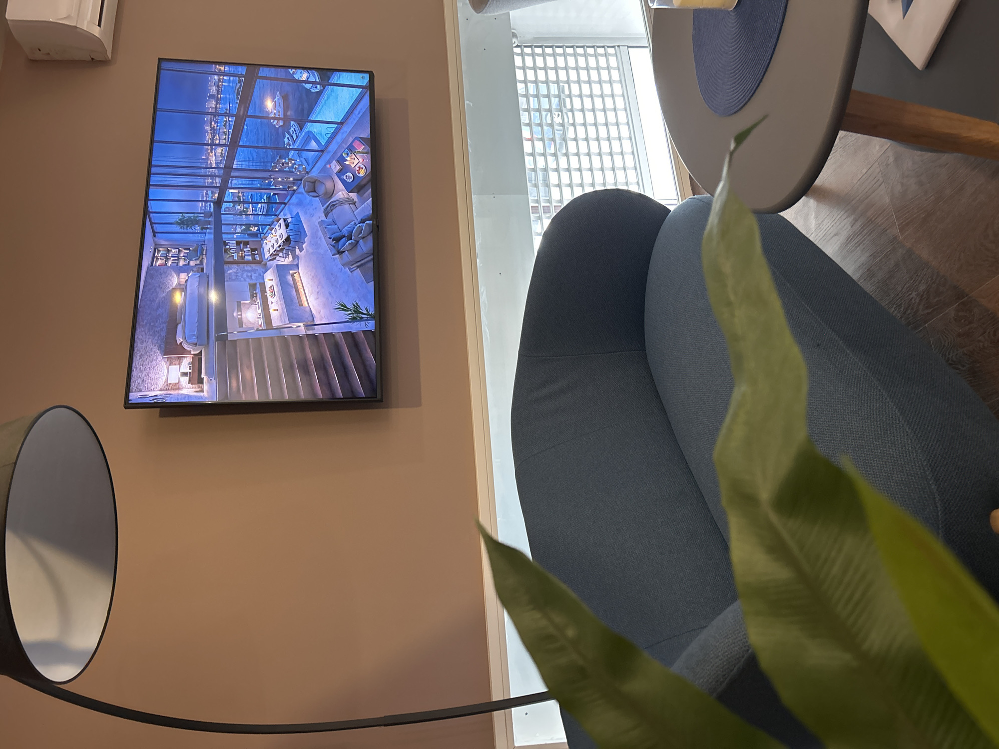 The image shows an interior scene with a focus on a seating area. A blue-grey armchair is partially visible, with a green plant in the foreground obscuring part of the view. A round table with a grey top and a blue center sits next to the chair. On the wall, there is a flat-screen television displaying an image of a luxurious interior space with a view of what appears to be a waterfront or coastal area. To the left of the television, a floor lamp with a circular shade is positioned. The wall is painted in a warm, muted beige or tan color. A window with a grid-like pattern is located to the right of the television. The location of the scene is Unknown, Unknown.