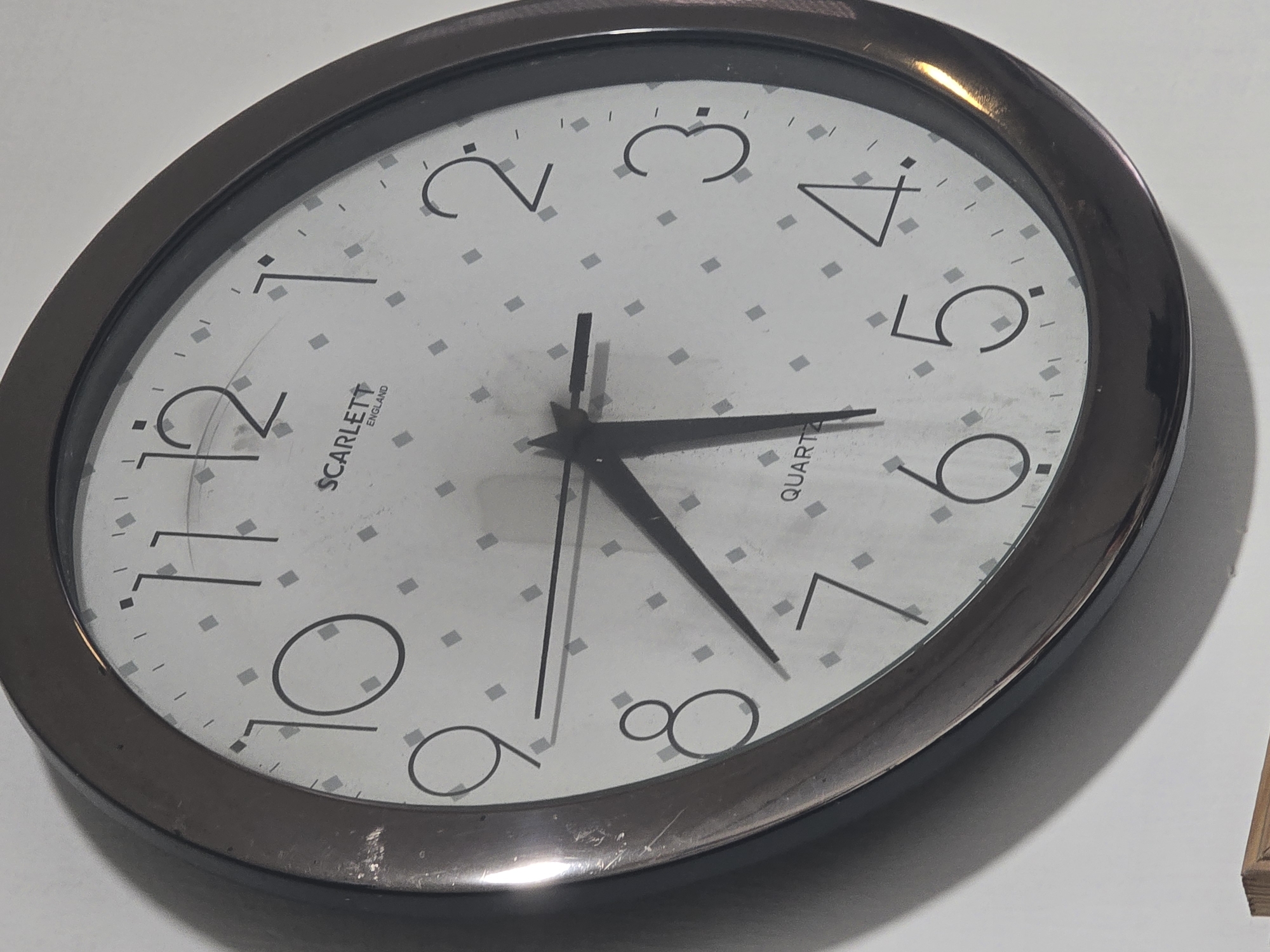 This is a close-up, angled shot of a round, analog wall clock. The clock face is white with a subtle diamond pattern and has black numbers indicating the hours from 1 to 12. The hour hand is pointing between the 4 and 5, closer to the 5. The minute hand is pointing directly at the 7. There is also a thin, black second hand that is pointing directly at the 11. The clock has a dark, possibly metallic, bezel. The clock is mounted on a plain white wall. There is a shadow cast on the wall behind the clock due to the angle of the shot. There are no people or any other objects visible in the frame. The location is not specified within the image itself.