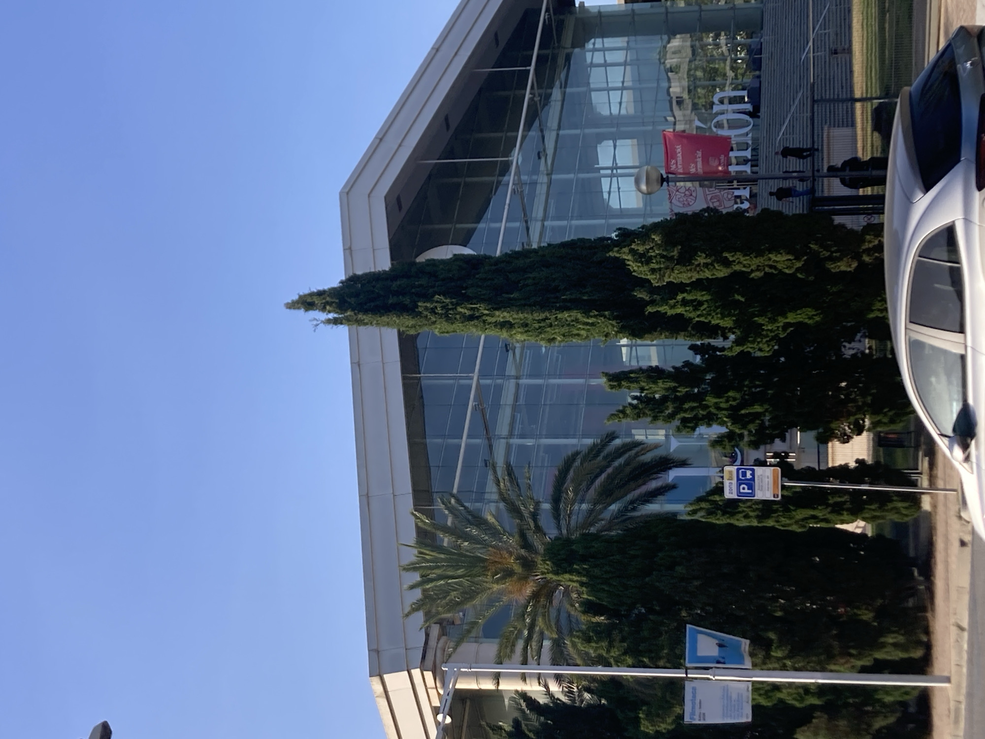 The image depicts an outdoor scene with a modern glass-fronted building. Several trees, including a tall conifer and a palm tree, are in the foreground, partially obscuring the building's facade. A white car is parked on the right side of the frame. The sky is clear and blue, indicating it is daytime with good weather. The building features large glass panels that reflect the surroundings. A red banner is visible hanging from a pole in front of the building. There are a couple of signs on poles, one of which appears to be a parking sign. Some people are visible walking near the building entrance, but they are small in the frame and not clearly identifiable. The overall impression is that of a bright, sunny day in an urban or commercial area, possibly near an event or exhibition given the banner. Some text is visible on the banner and signs, but it is not legible due to the image resolution and orientation. The location context of Barcelona, Spain, suggests a Mediterranean climate.