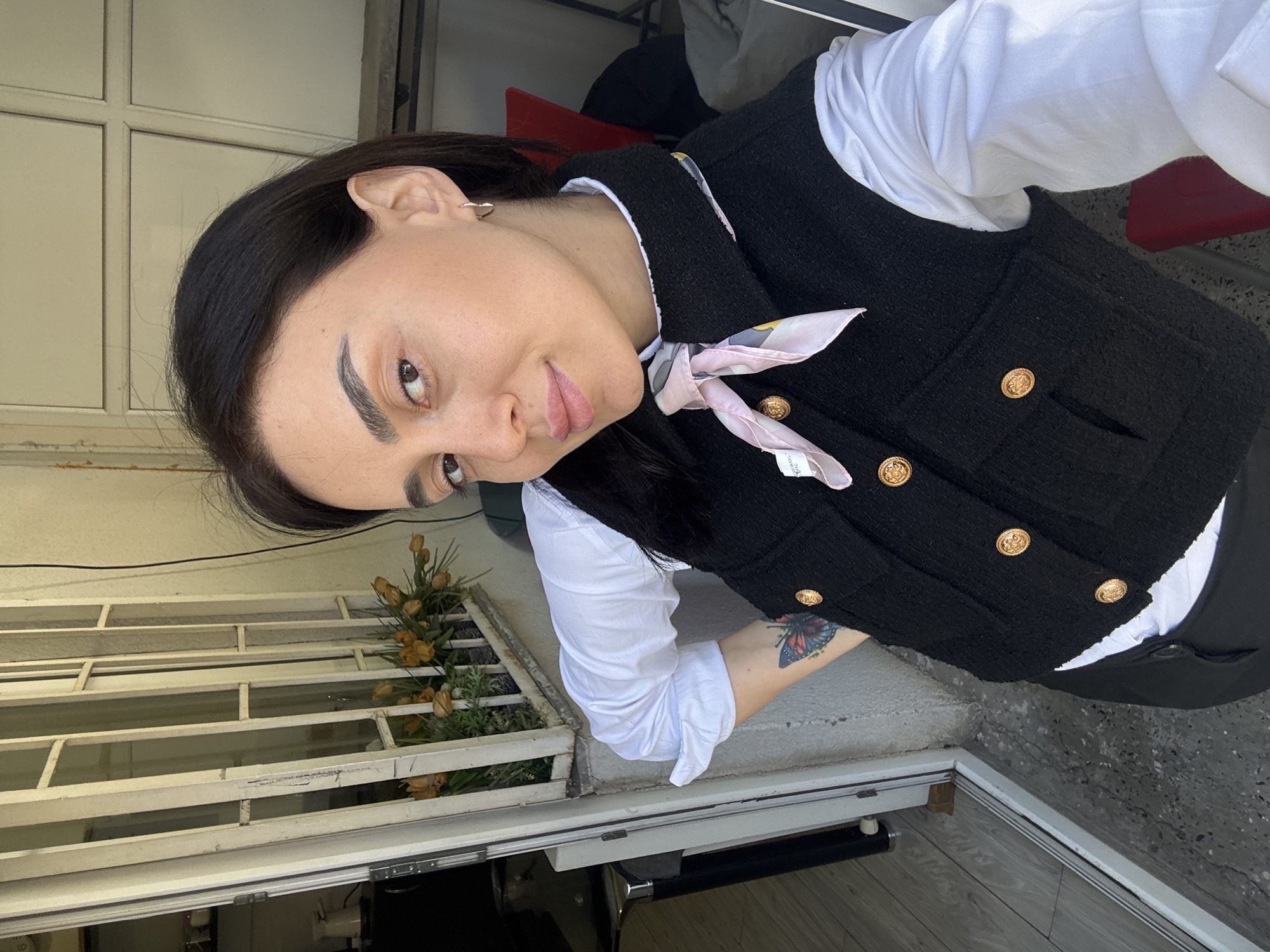 A woman is featured prominently in a self-portrait, looking directly at the camera with a slight, calm smile. She has dark hair, dark eyes, and well-groomed eyebrows. She is wearing a white long-sleeved collared shirt beneath a black, textured sleeveless vest adorned with multiple ornate gold buttons down the front. A light pink patterned silk scarf is tied around her neck. A colorful butterfly tattoo is visible on her left forearm. A simple silver hoop earring can be seen in her right ear.
The setting appears to be an indoor or enclosed outdoor space, such as a doorway or balcony, in what is identified as Tbilisi, Georgia. In the background to her right, there is a light-colored window frame or security gate with multiple rectangular panes and bars. Behind this, a plant with brownish-yellow, possibly dried or artificial, flowers is visible. A hint of a red chair can be seen in the upper right background. The ground in the foreground consists of a textured, grey concrete-like surface, leading to a darker area and what appears to be a door threshold or frame.
The woman is the primary subject, engaged in posing for the photograph. The time of day appears to be daylight, though no strong shadows are present, suggesting possibly an overcast day or indirect light. A small, illegible textile label is visible on her scarf. The overall mood conveyed by her expression is gentle and composed.