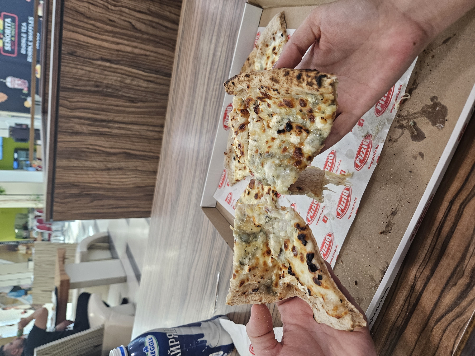 In Stara Zagora, Bulgaria, a close-up shot captures two hands holding slices of pizza over an open pizza box. The pizza appears to be a four-cheese variety with a golden-brown crust. The box, lined with paper featuring the "Pizzio" logo and website, rests on a wooden table. To the left, a bottle of milk with a blue and white label is visible. In the blurred background, a person is seated at a table, drinking from a bottle. The overall environment suggests a casual dining setting, possibly a food court or restaurant within a shopping mall, given the presence of wooden paneling and distant seating areas with red chairs.