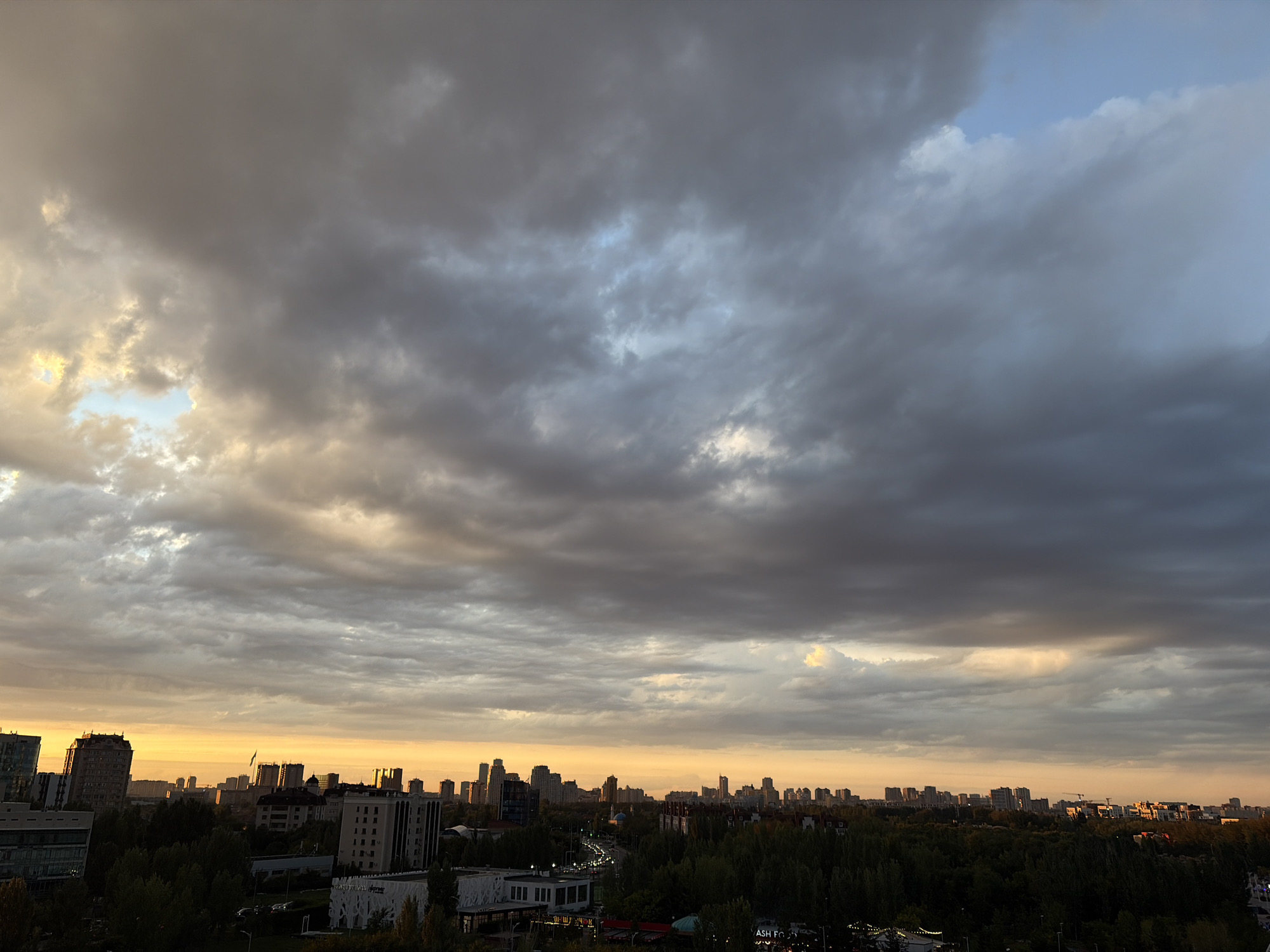The image depicts a panoramic view of the cityscape of Astana, Kazakhstan, during what appears to be sunset or sunrise. The sky is dominated by dramatic, heavy clouds, with patches of light breaking through, casting a warm, orange glow on the horizon. Below the sky, a dense urban landscape unfolds, characterized by a variety of modern and older buildings, including high-rise structures and lower residential or commercial buildings. A lush green canopy of trees separates the city's skyline from the foreground. Roads and some vehicles are visible, suggesting ongoing urban activity. The overall atmosphere is one of a transitioning sky over a bustling city, with a strong emphasis on the interplay of light and shadow created by the clouds.