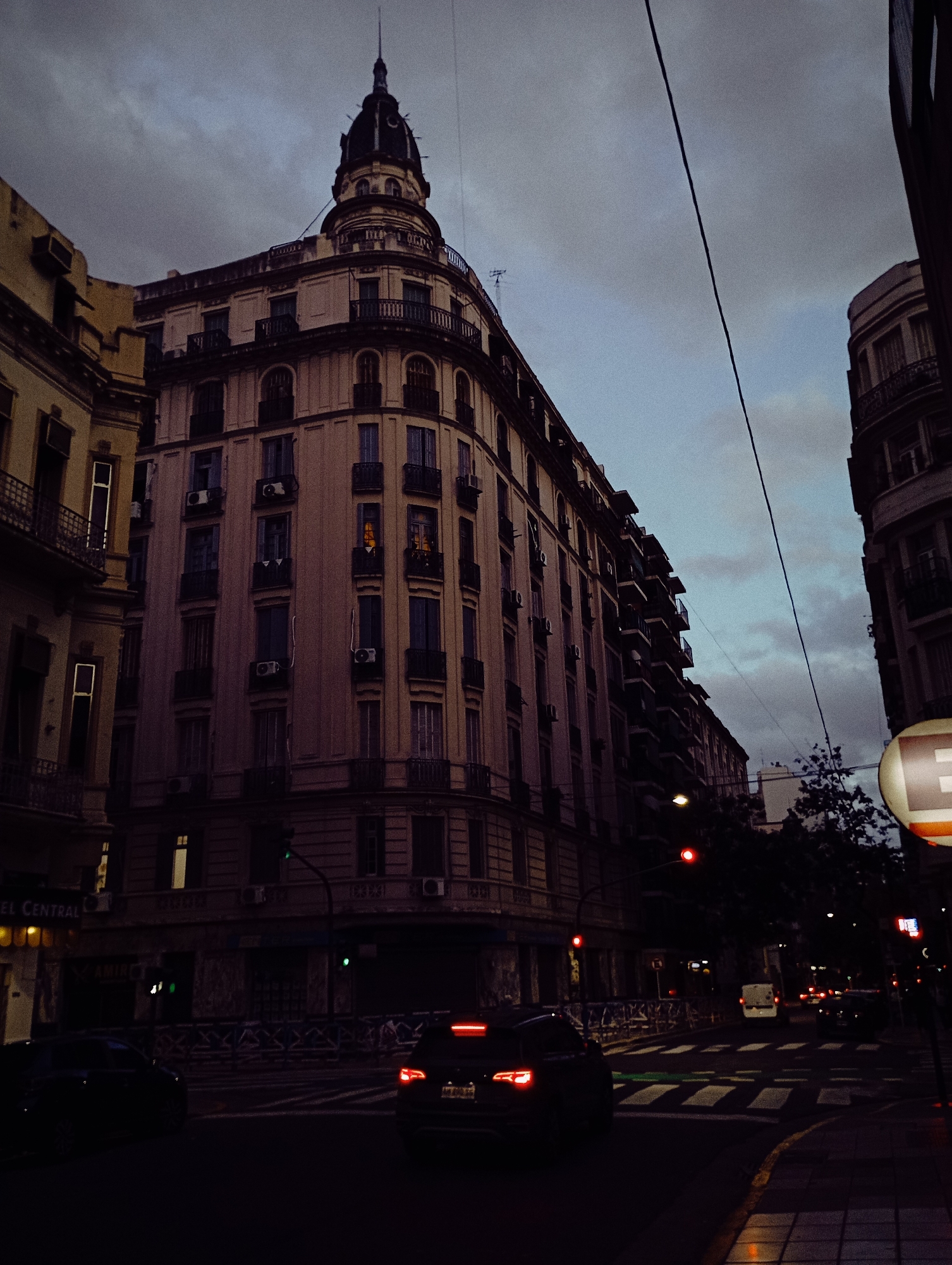 The image shows a street scene in Buenos Aires, Argentina, at dusk. The main focus is a corner building with a distinctive dome and multiple floors of balconies and windows, many with air conditioning units. The building's facade is a light color. Streetlights and car headlights illuminate the scene. A vehicle with illuminated tail lights is visible in the foreground, crossing the street, which has a green crosswalk. Other cars are on the street as well. The sky is overcast. A utility line stretches across the upper portion of the image. A building is partially visible to the left, and a sign is partially visible on the right.