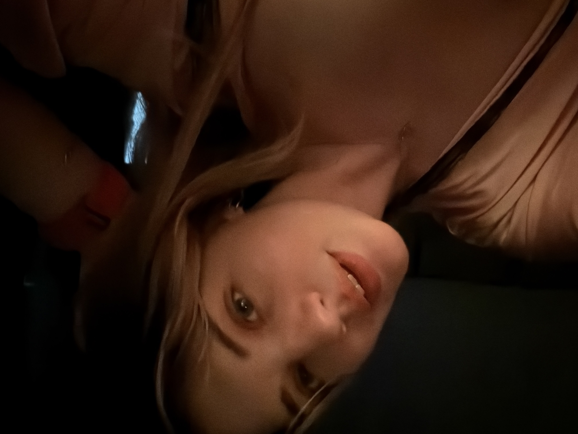 The image shows a woman with light skin and blonde hair. She is wearing a light pink top with a dark strap. Her face is angled, with her eyes open and looking slightly to the left. Her lips are slightly parted. The background is dark. There is no information to suggest that the image was taken in the city of Большая Шильна, Russia.