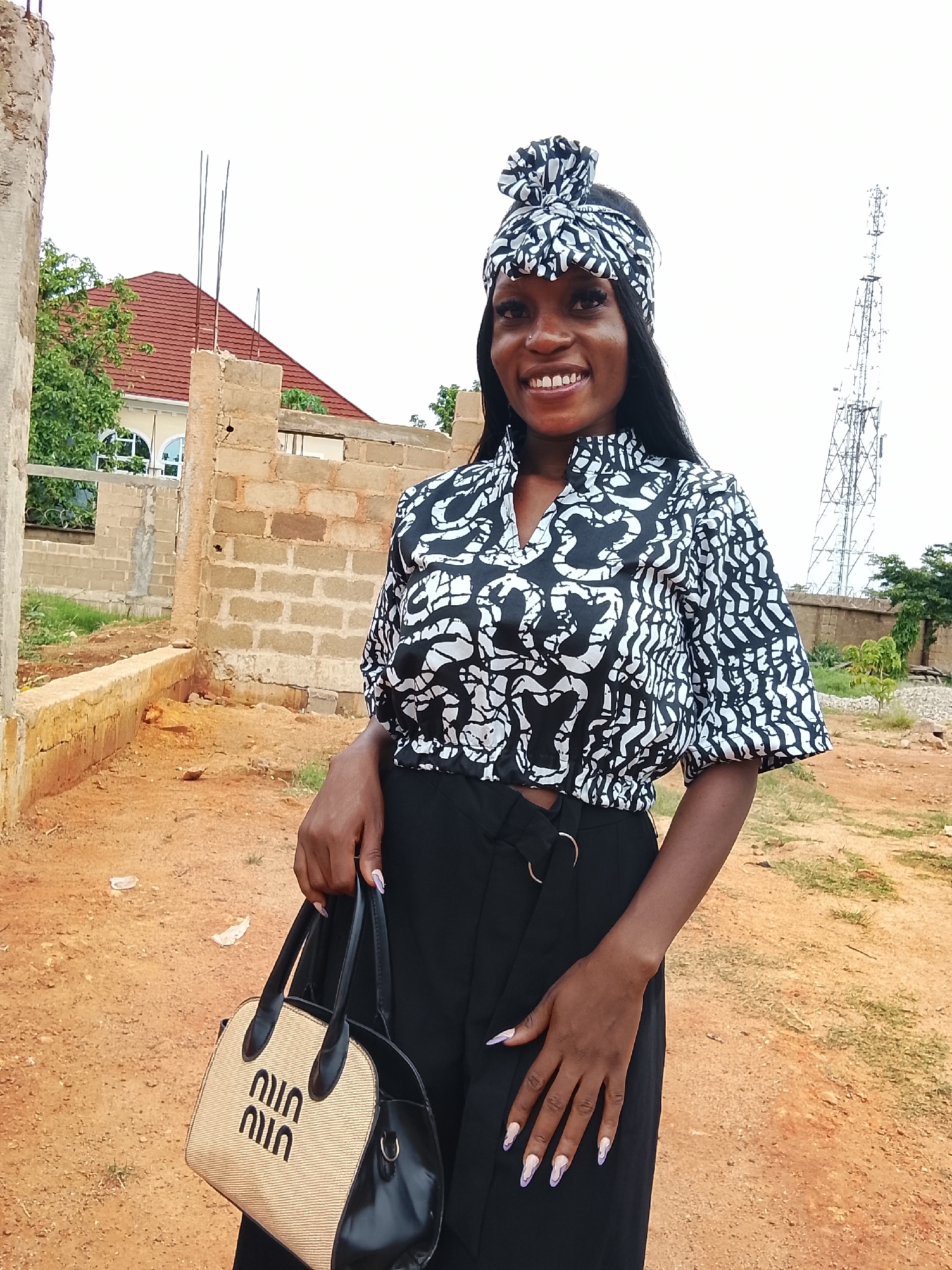 A young woman stands outdoors holding a handbag. She is wearing a black-and-white patterned top and a matching headband, along with black pants. She is smiling and looking toward the camera. The bag she's holding has the logo "MIU MIU" printed on it. Behind her is a partially constructed brick building, and in the distance is a communication tower. The ground is reddish-brown dirt. Based on the presence of the brick building under construction, and the overall development level, the location is likely in a developing area within Barikin Sabon Gidan Kanat, Nigeria.